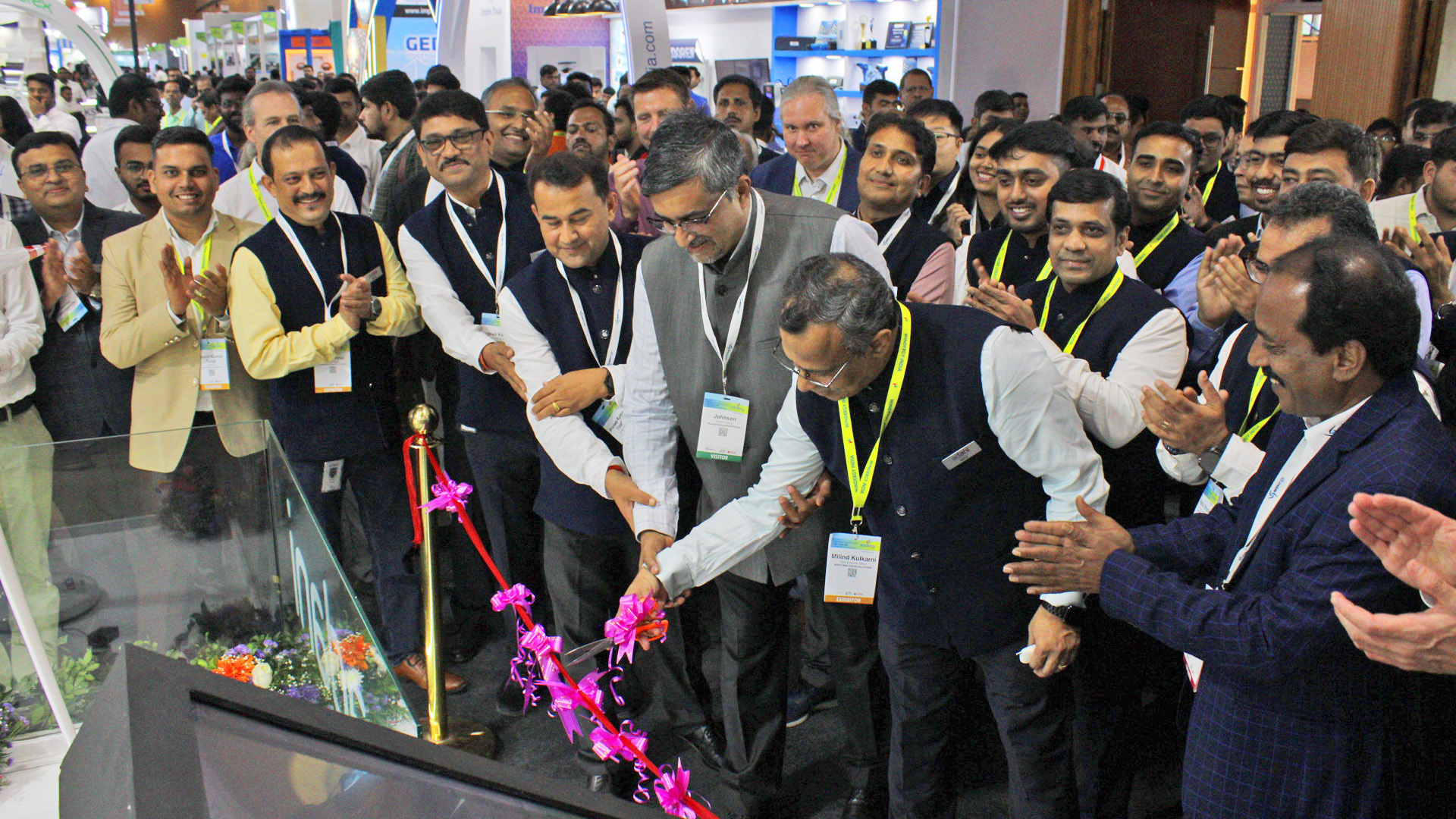 Two Indian men cut a pink ribbon, with other men clapping at an event.