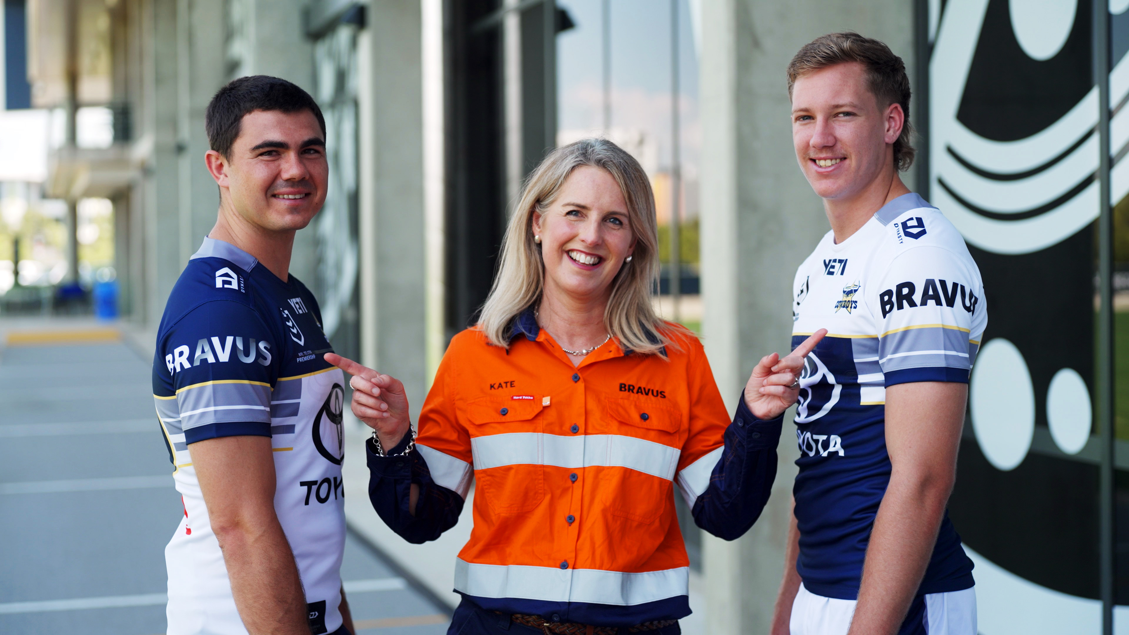 Smiling woman in orange work shirt points at two men in sports jerseys, who also point at her.