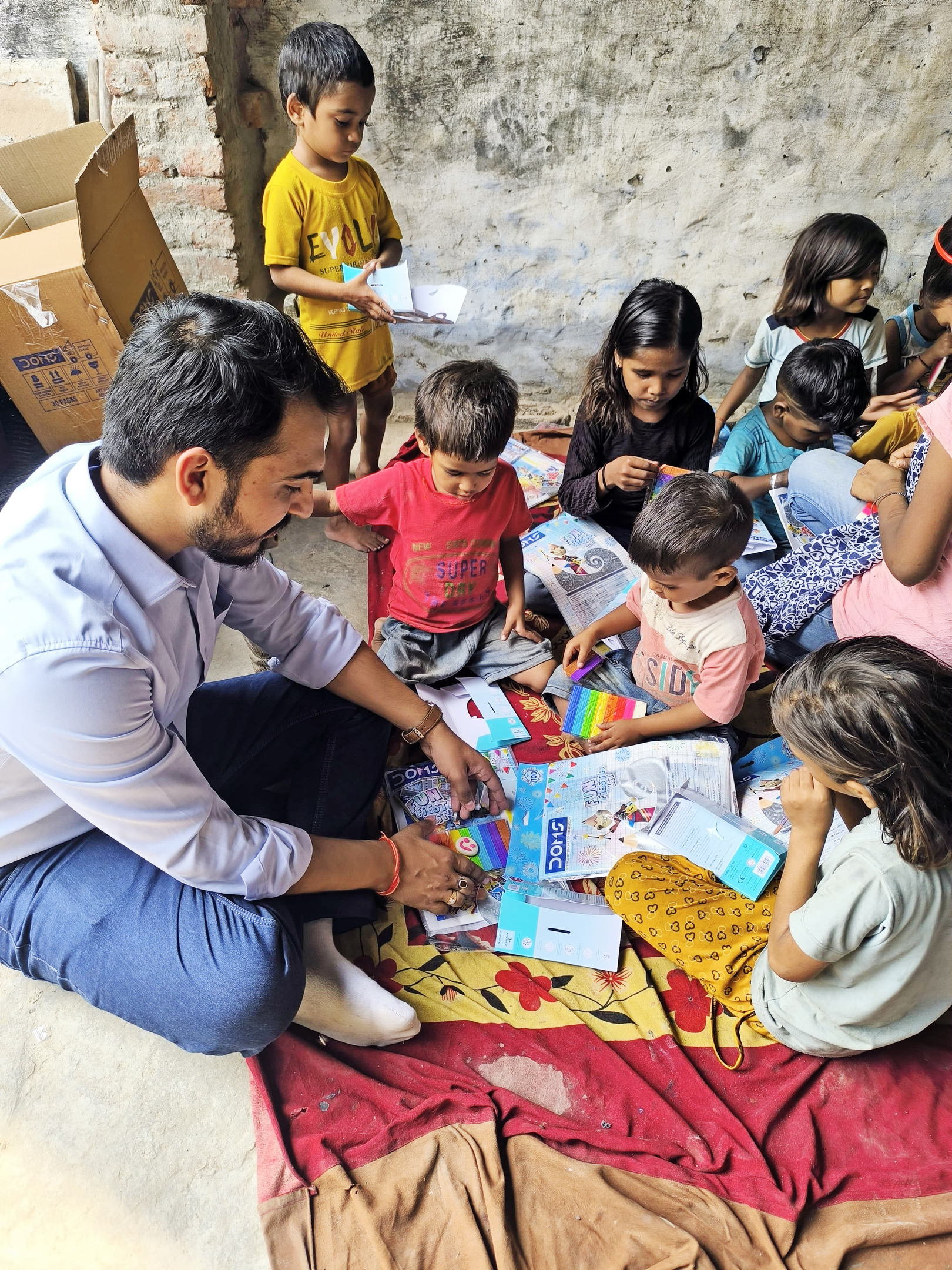 A man sits with children, sharing school supplies and art materials.