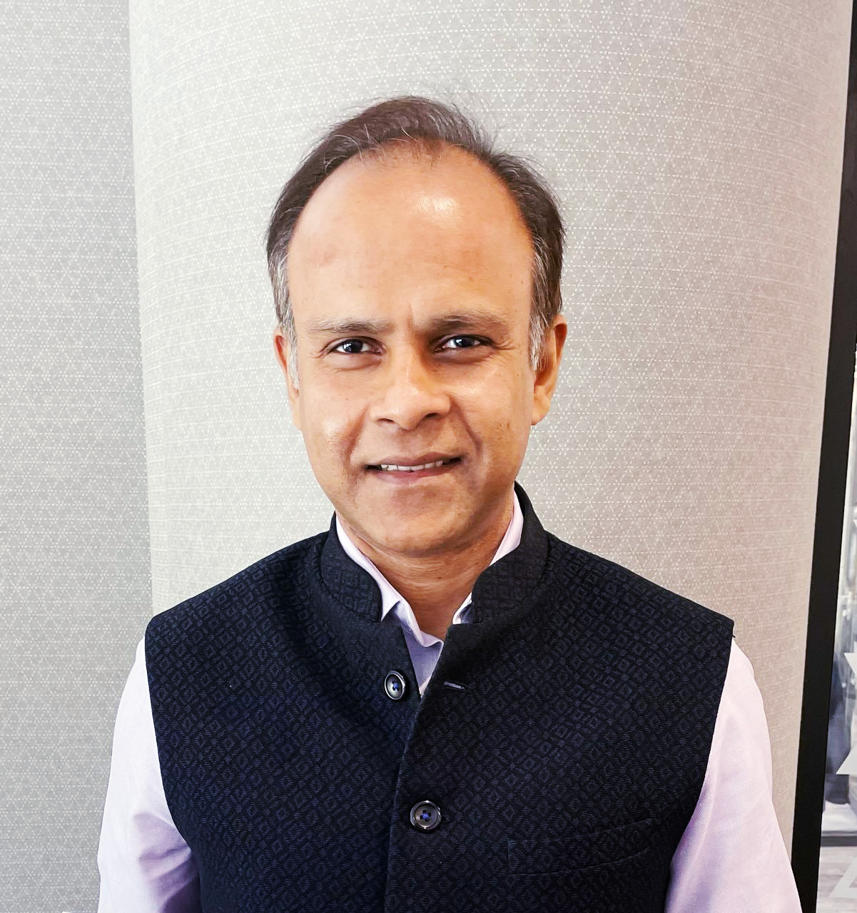 Smiling man in a dark patterned vest over a light shirt.