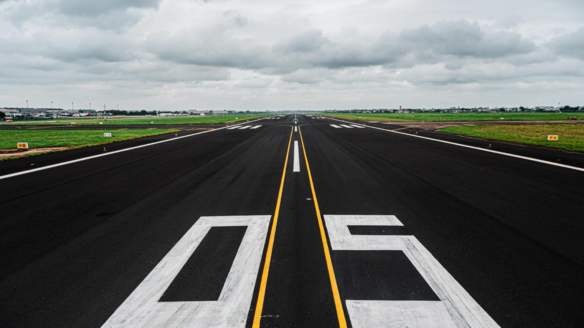 Airport runway with '05' marking, yellow and white lines, stretching to the horizon under a cloudy sky.