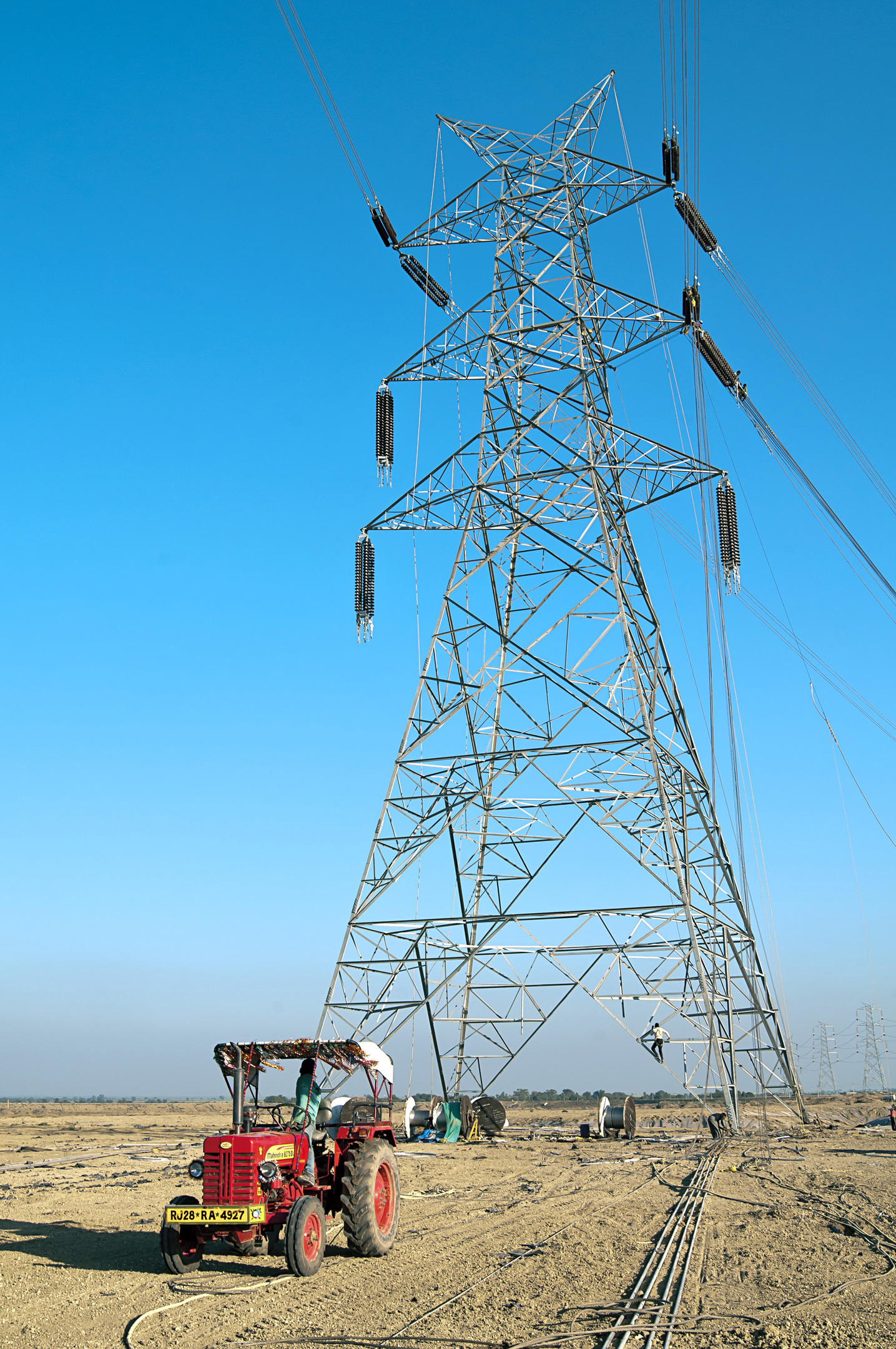Workers on a tall electricity pylon, with a red tractor and cables in a dry field under blue sky.