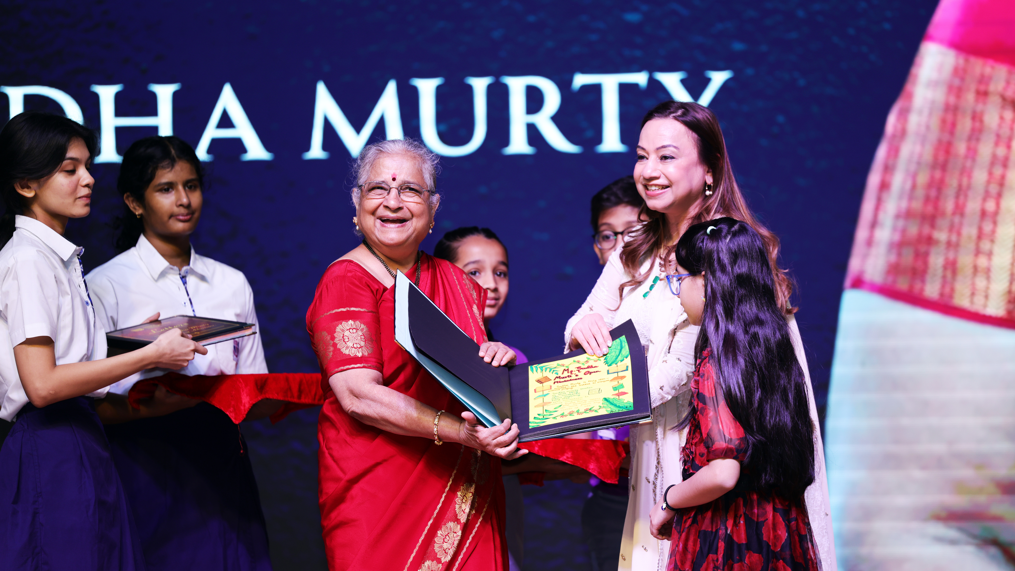 Sudha Murty and another woman smiling and holding an award on stage with children in the background.