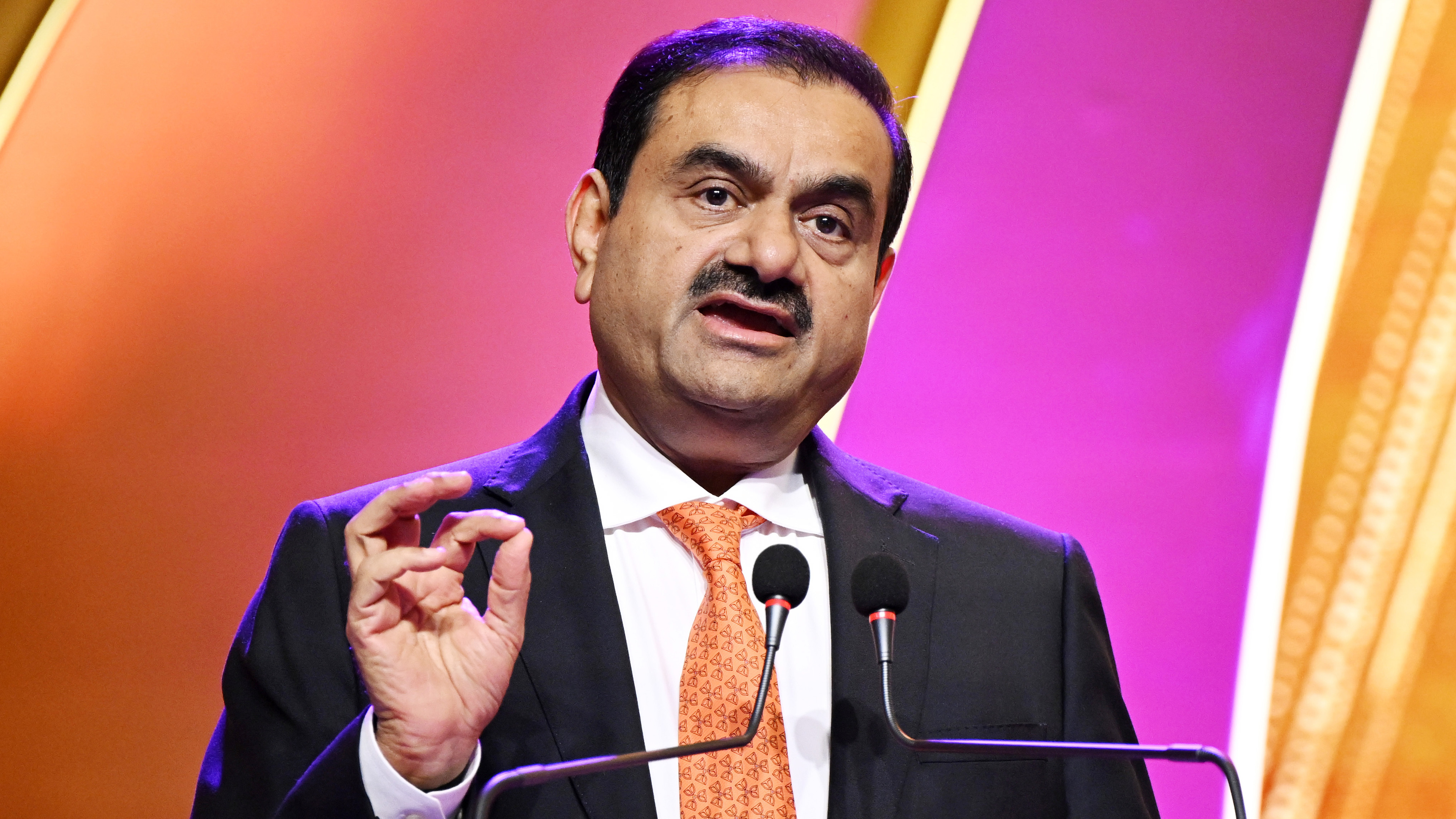 Gautam Adani speaks at a podium, gesturing, vibrant orange and pink background.