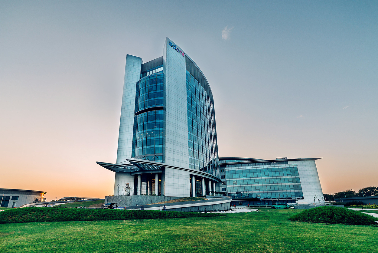 Modern Adani building with glass architecture, green lawn, and sunset sky.