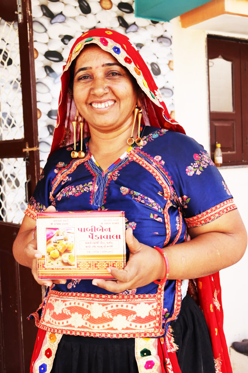 Smiling Indian woman in colorful traditional attire holding a box of Pedawala sweets.