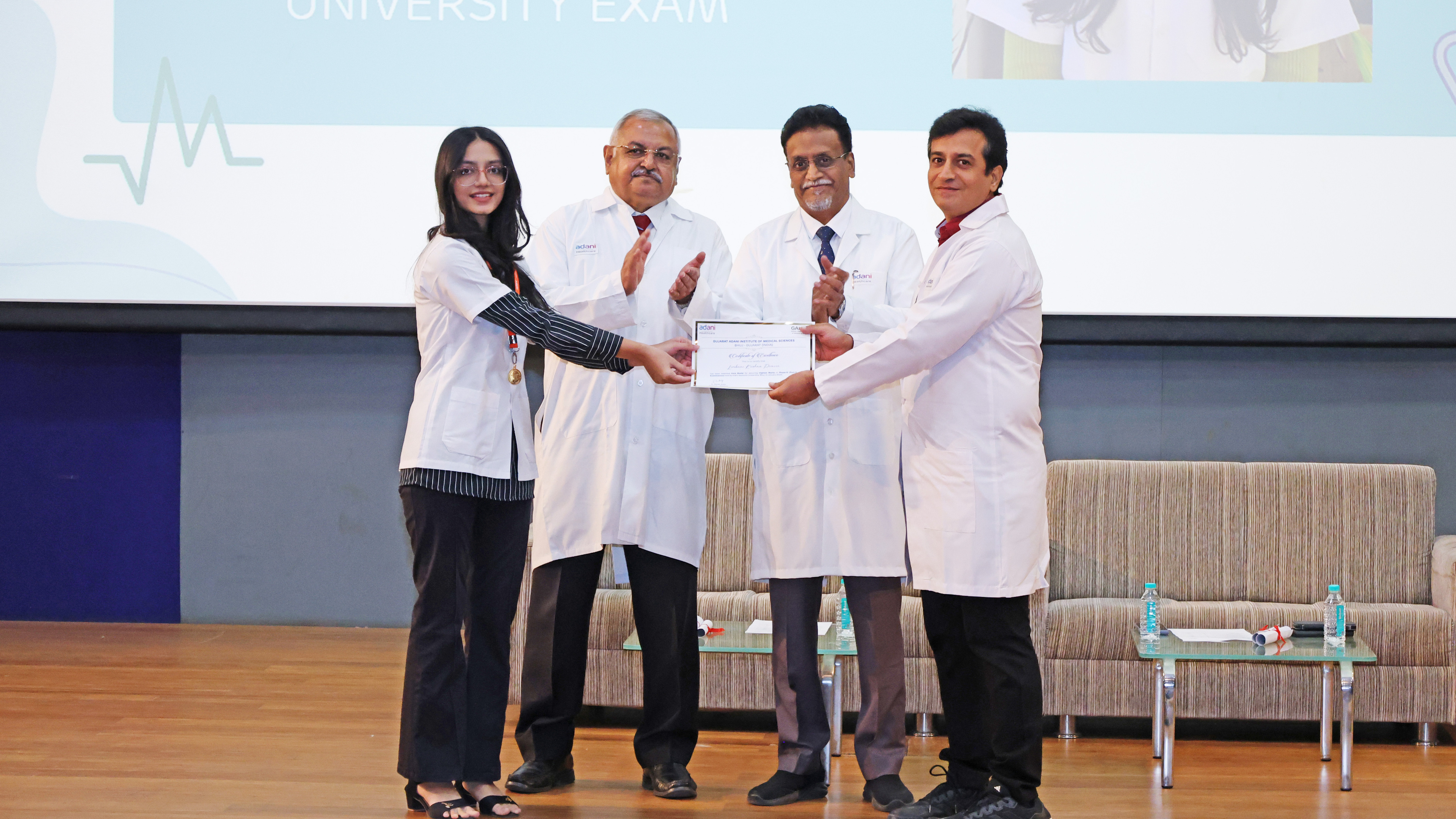 Woman in lab coat receives certificate from two men on stage, another man claps.