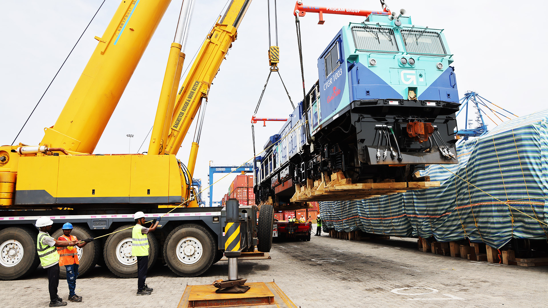 Giant crane lifting a blue train locomotive at a port, with workers observing.