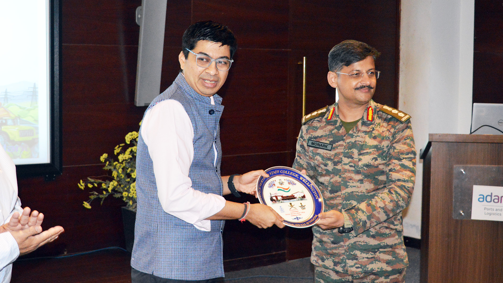 A man in civilian clothes hands a Staff College, Wellington plaque to military officer Monark.