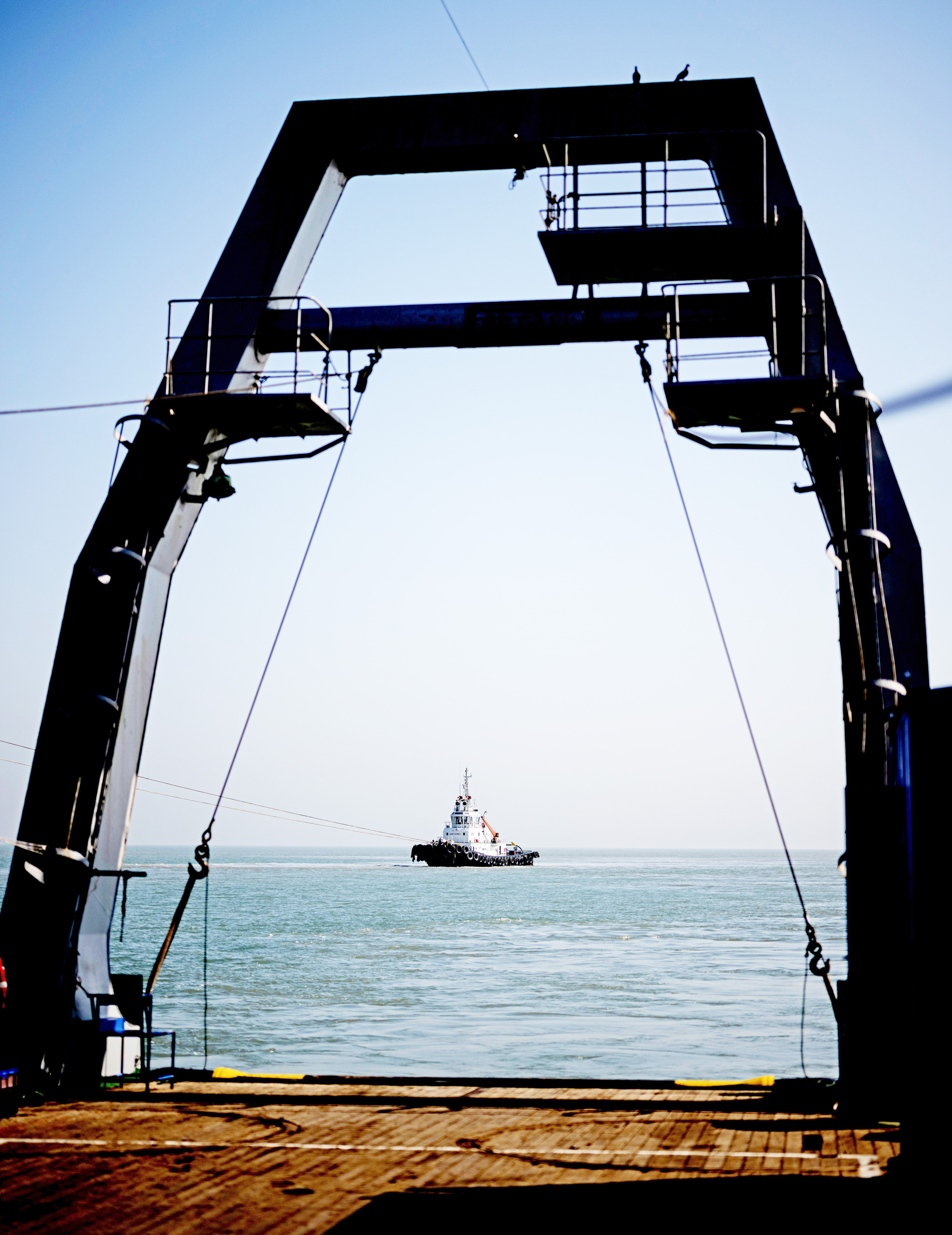 A dark, arch-like structure frames a distant tugboat on the ocean, viewed from a wooden deck under a clear sky.