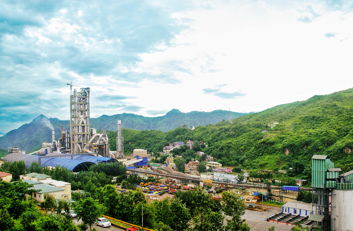 A cement factory and town nestled in a lush green mountain valley under a cloudy sky.