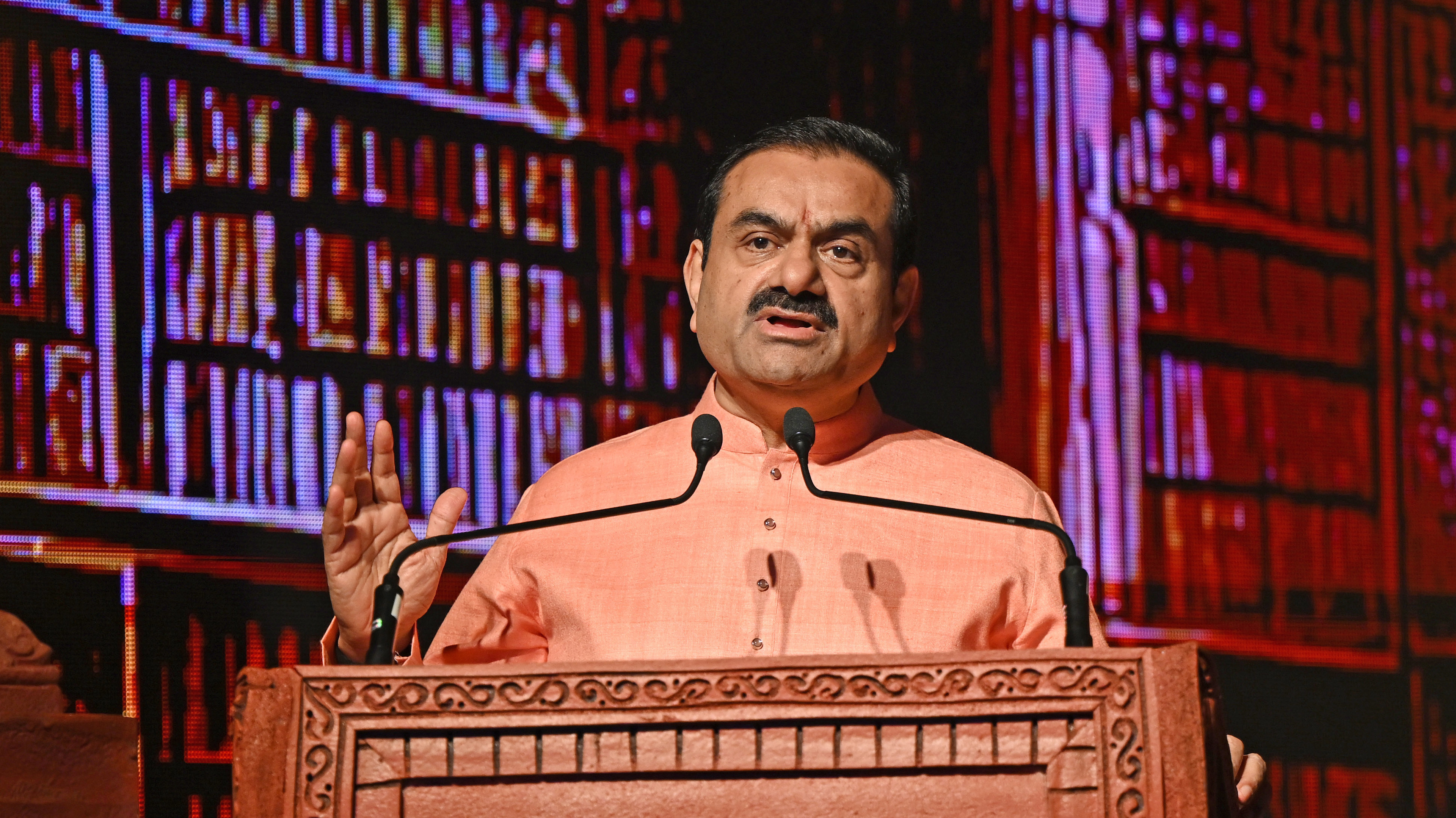 Man in peach kurta speaks at a podium, hand raised, with a digital bookshelf background.