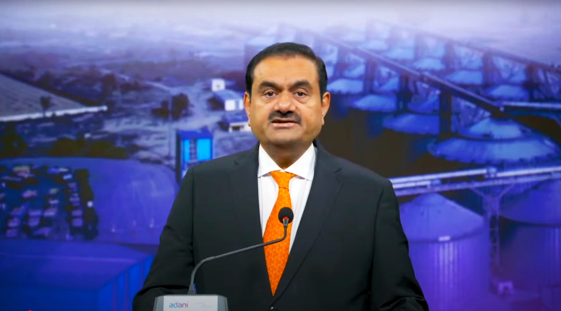 Gautam Adani speaking at a podium with industrial silos in the background.