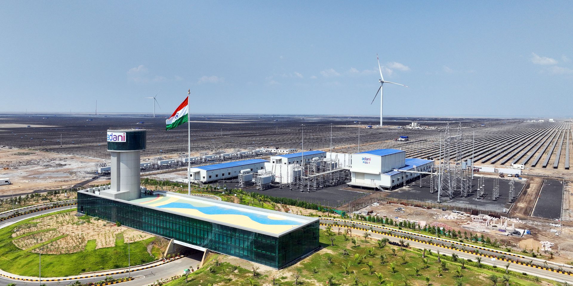 Adani renewable energy plant with vast solar fields, wind turbines, buildings, and Indian flag.