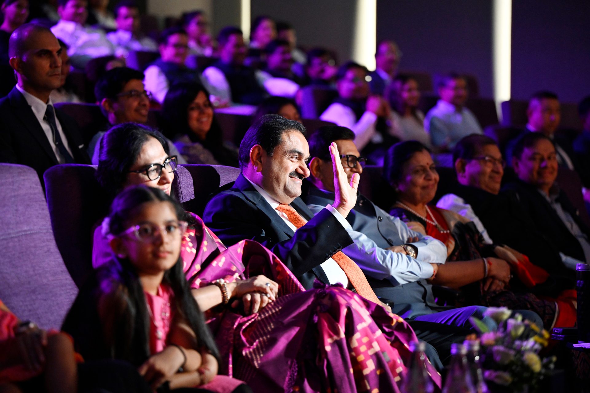 Gautam Adani smiles and waves to an audience in a hall, with a child in the foreground.