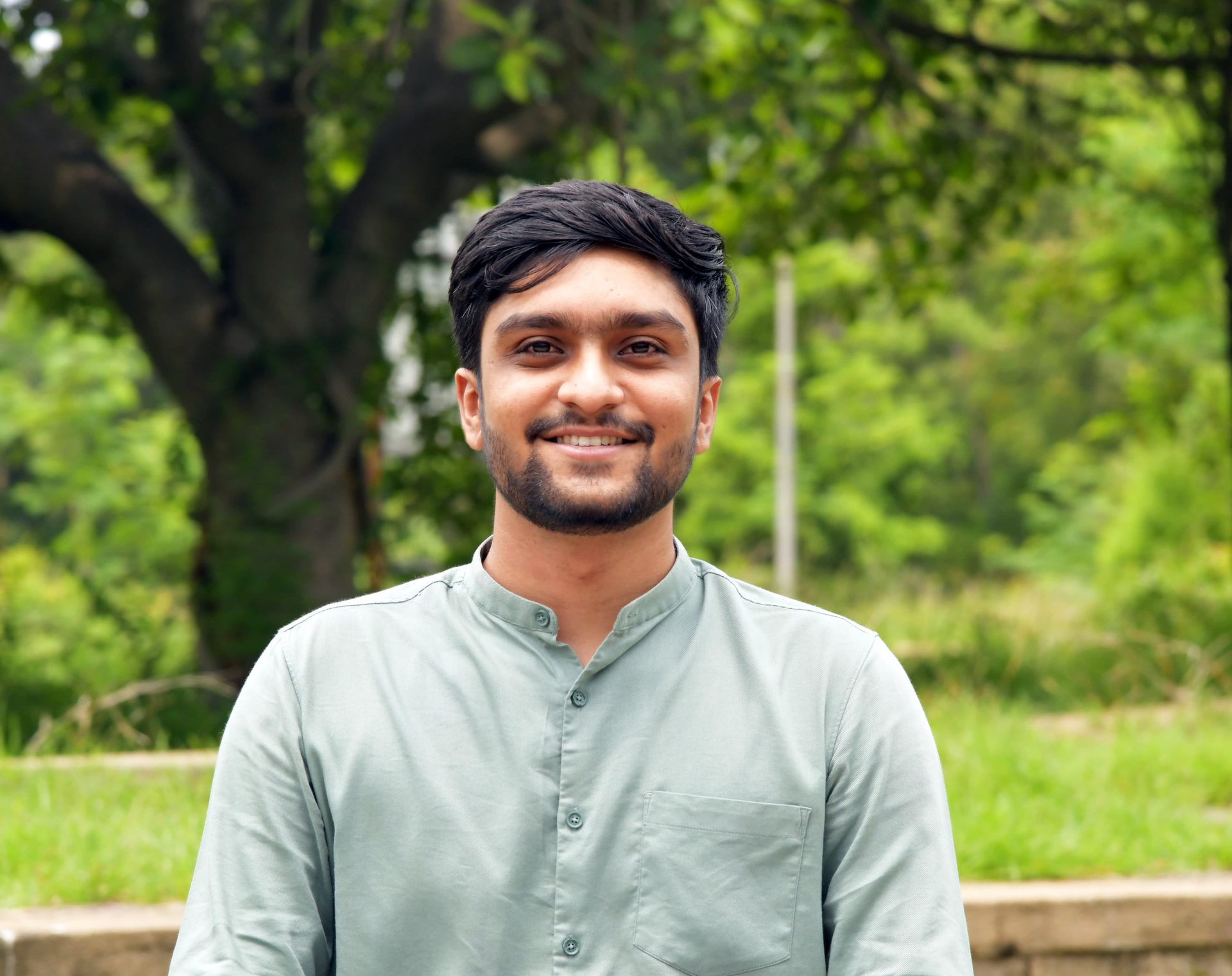 Smiling young man in light shirt, outdoors with green trees.