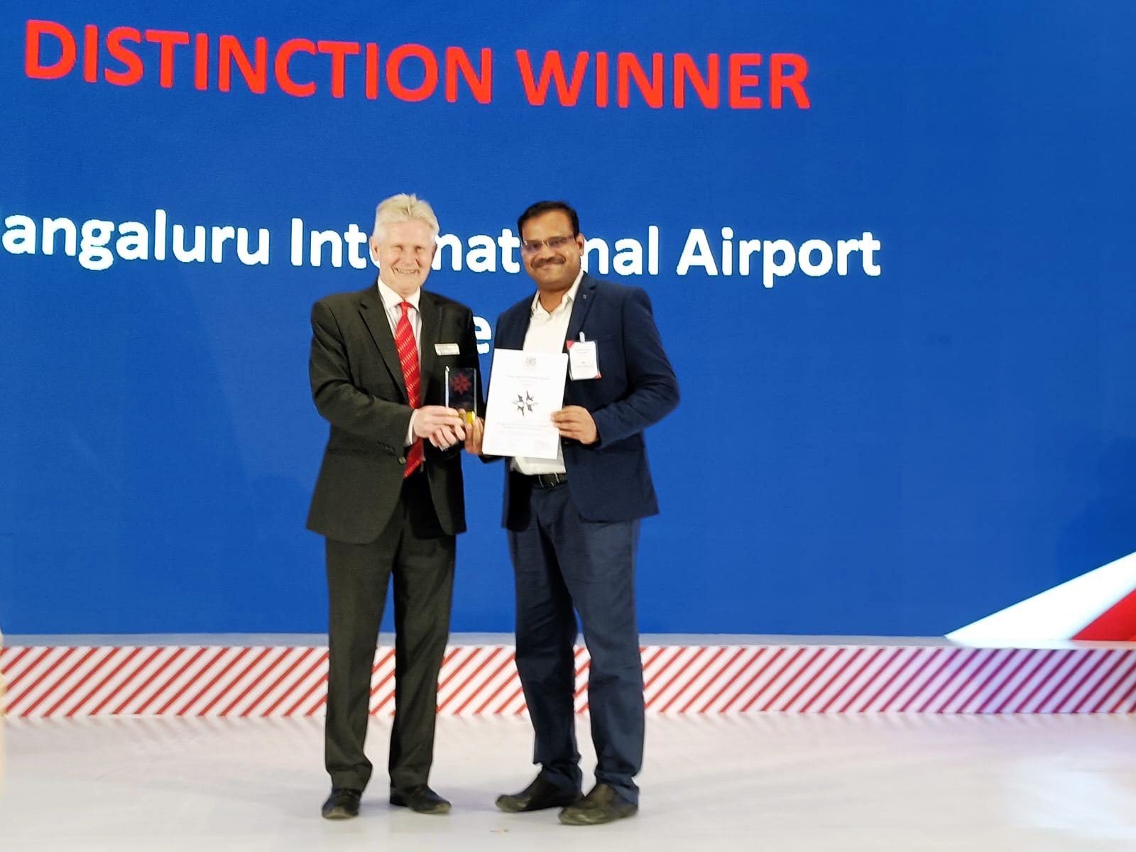 Man presents award to another. Screen: 'Distinction Winner, Bengaluru International Airport'.