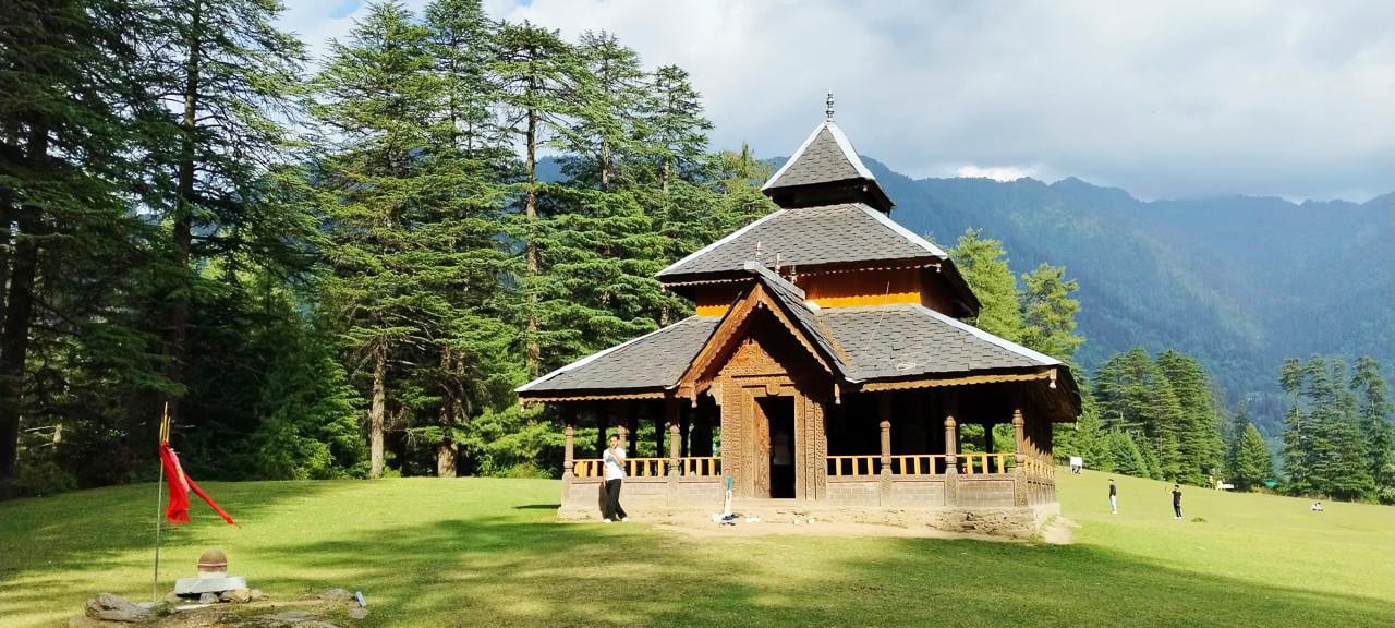 Wooden temple in a grassy field, surrounded by tall pine trees and mountains under a cloudy sky.