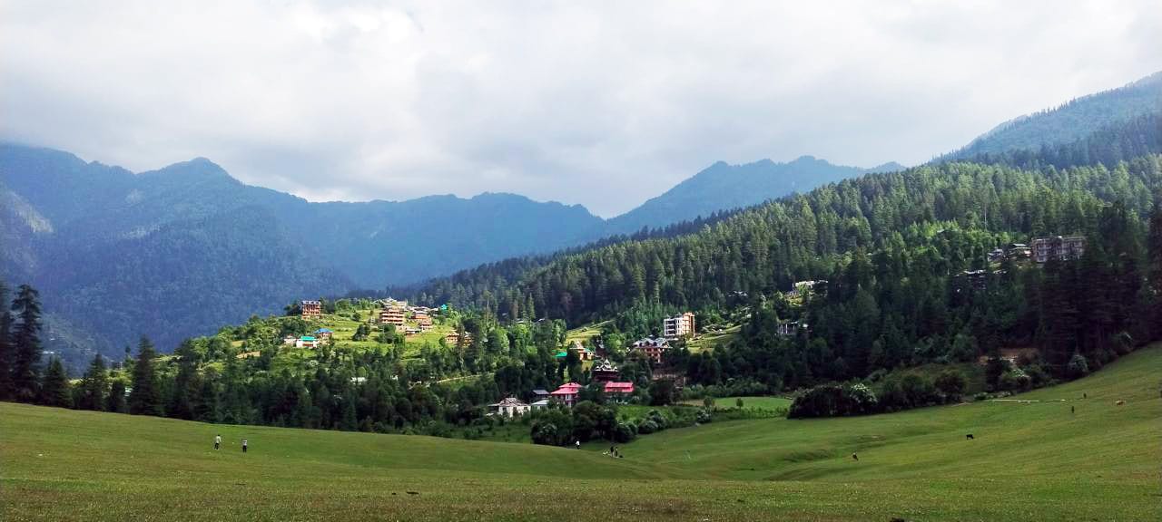 Lush green valley with a village nestled in dense forests, backed by mountains under a cloudy sky.