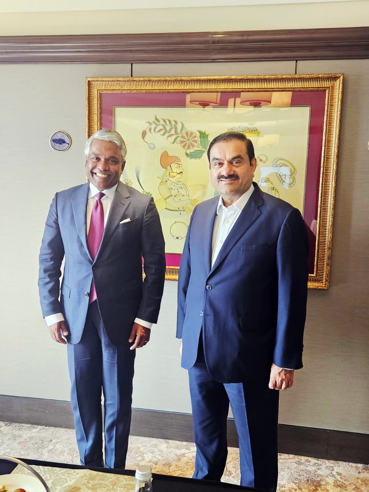 Two smiling men in suits, including Gautam Adani, stand side-by-side indoors.
