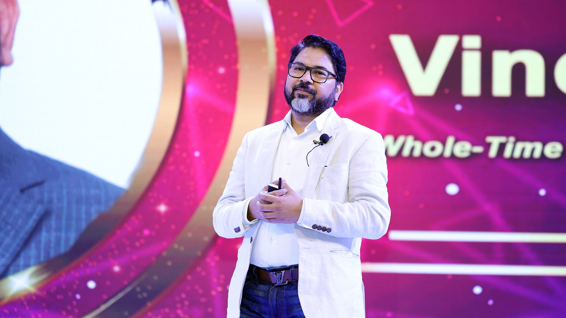 Man with glasses and beard in light jacket speaking on stage; screen behind shows "Vin Whole-Time".