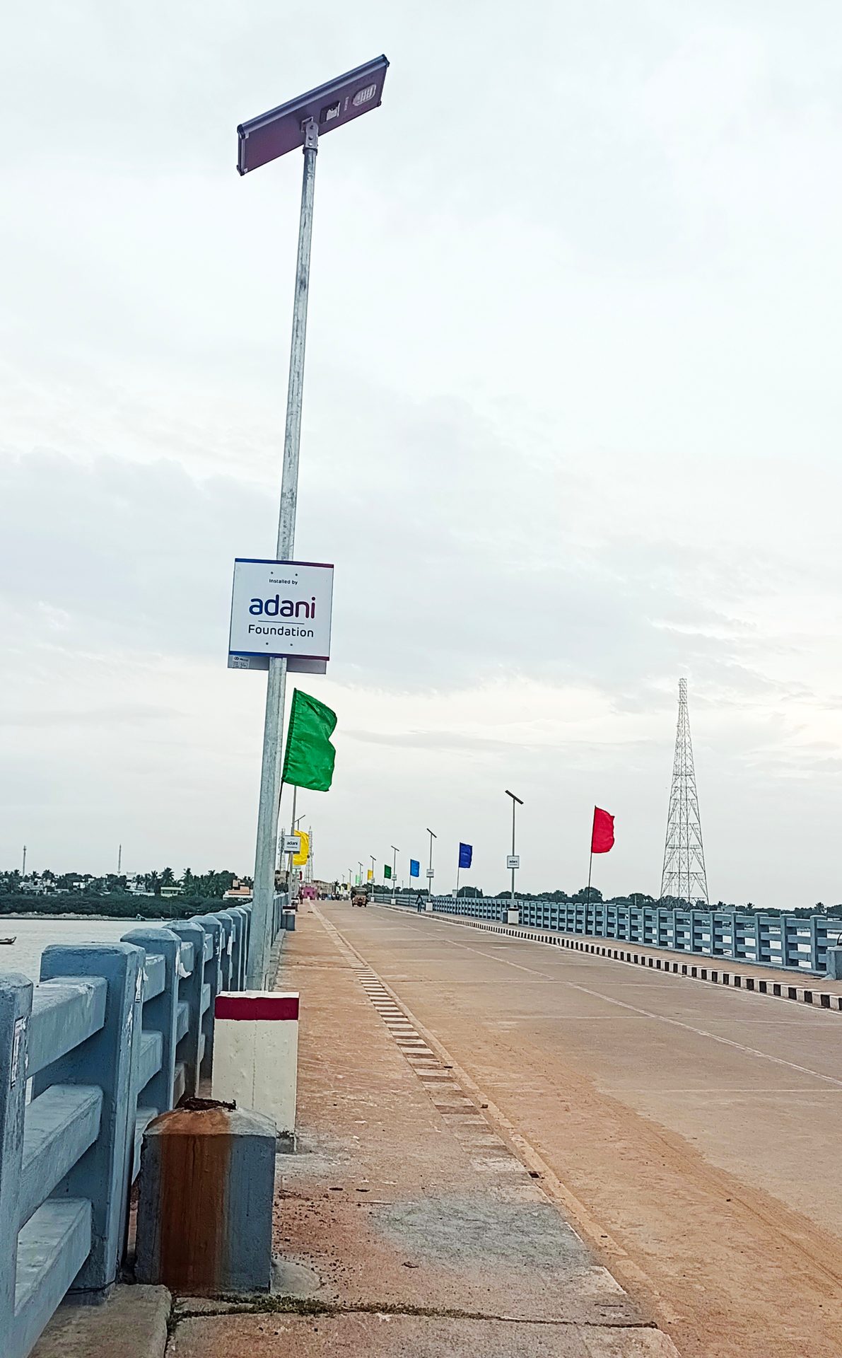 A bridge with solar streetlights, colorful flags, and an Adani Foundation sign, across water.