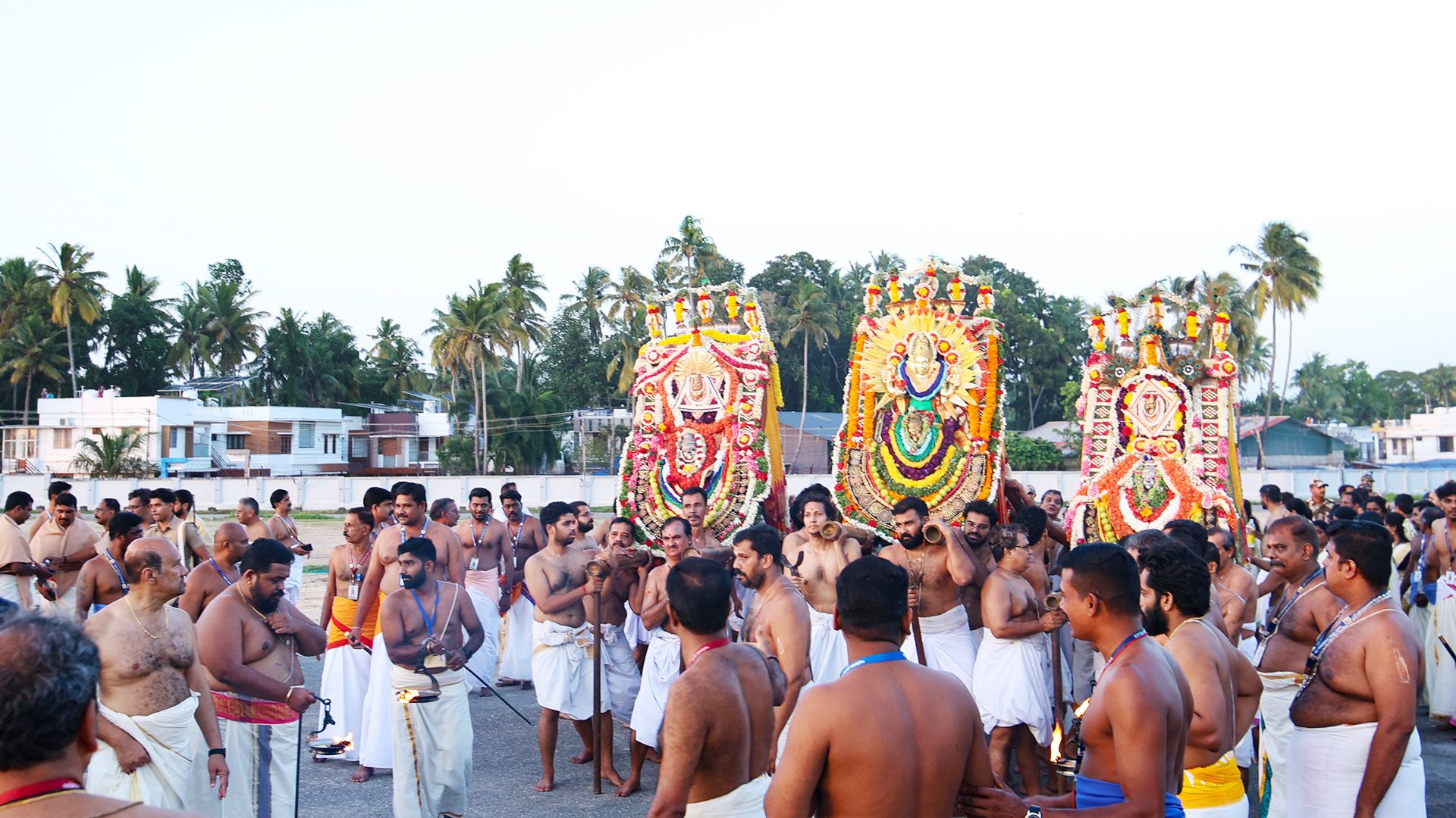 Bare-chested men in white dhotis carry ornate, colorful deity palanquins during a traditional festival.