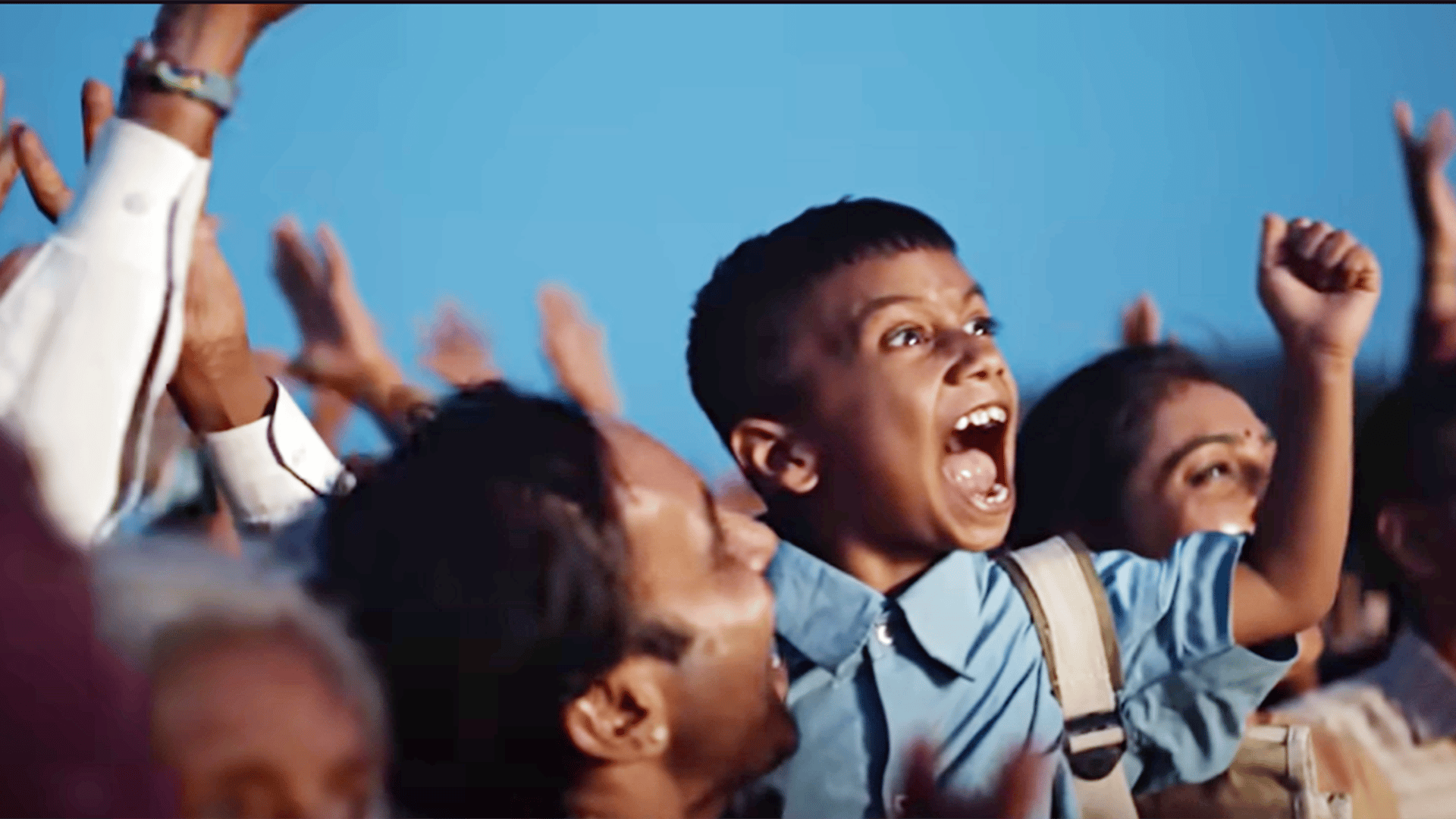 Excited boy shouts with raised fist in a cheering crowd against a blue sky.