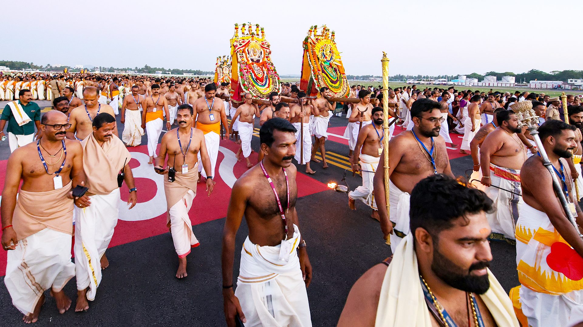 Men in traditional attire, some shirtless, carry ornate palanquins and lamps on an airport runway.