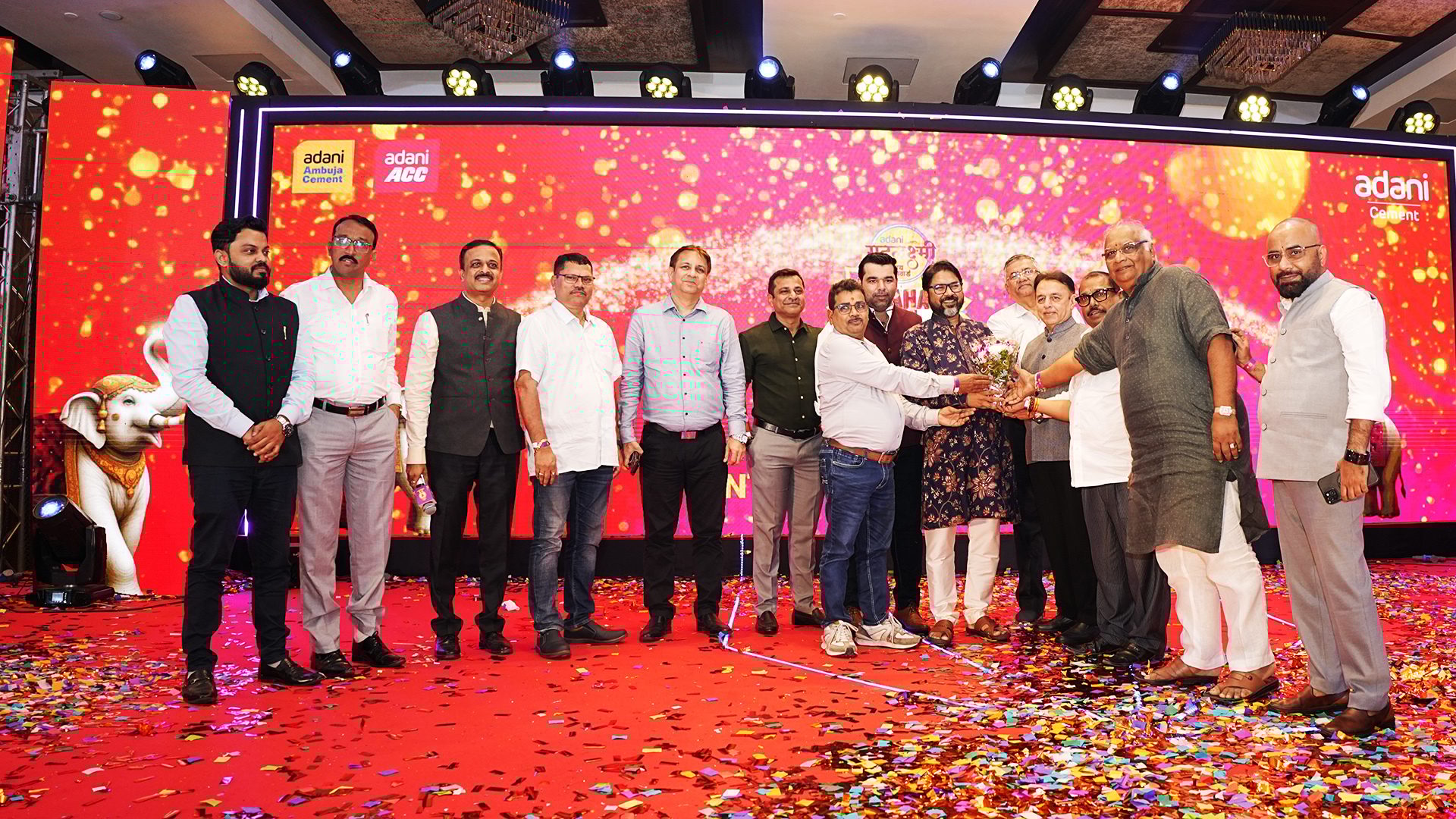 Group of men on a stage with Adani logos, one receiving flowers amidst confetti.