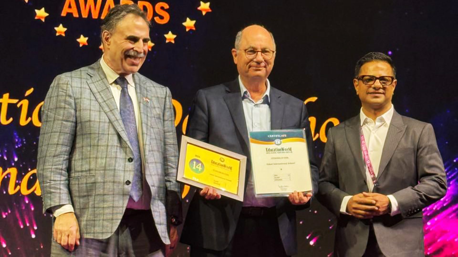 Three men in suits on stage, one holds two "EducationWorld" awards.