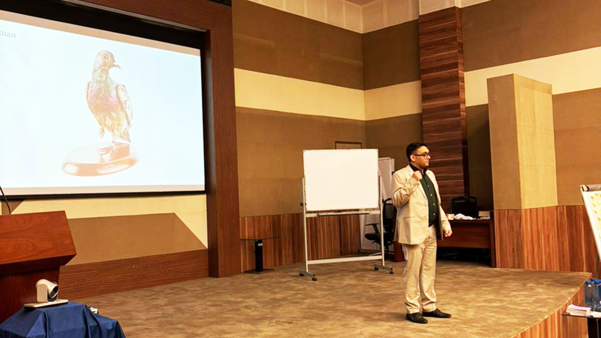 Man in a light suit presenting on a stage with a projector screen showing a bird image.