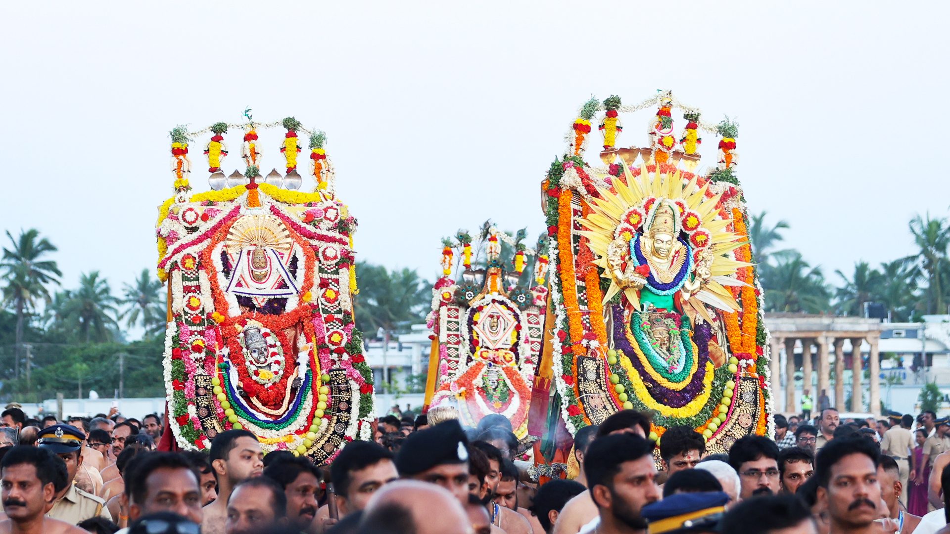 Three ornate, flower-decked chariots with deities carried by a large crowd during a religious procession.