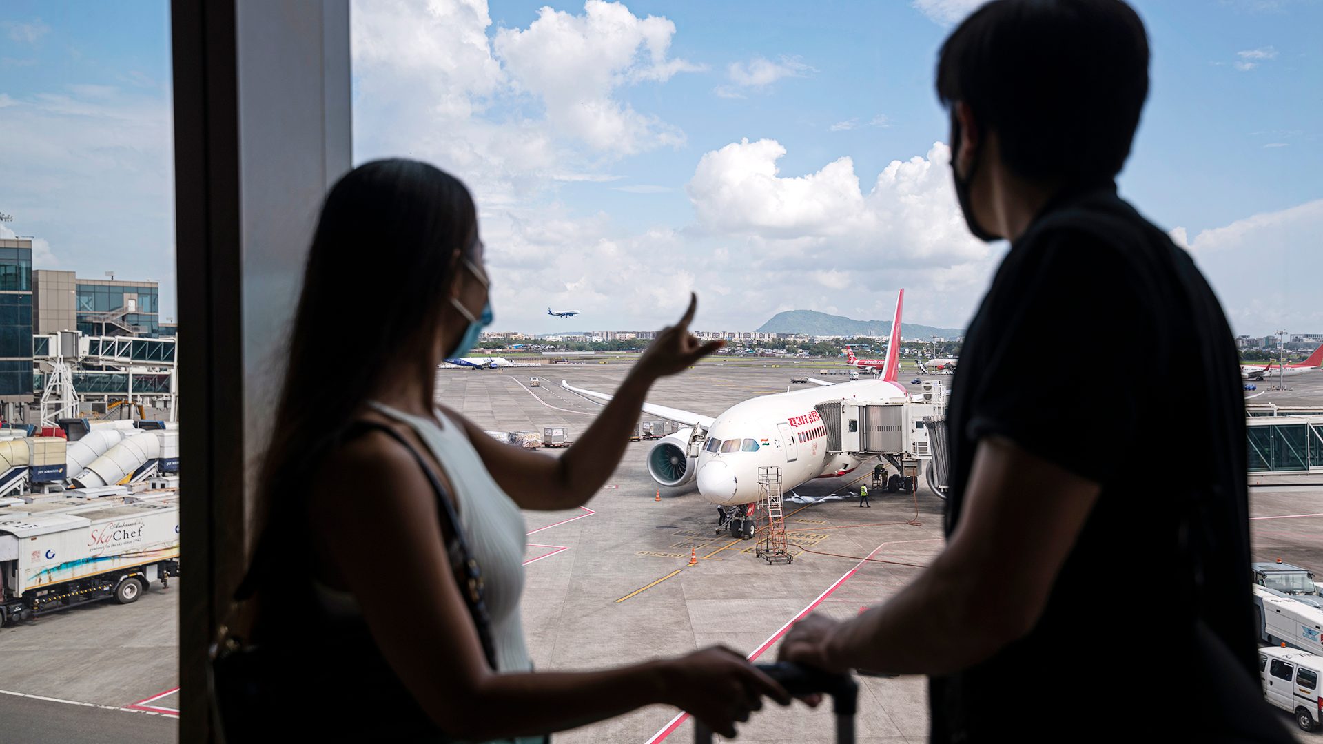 Two people look out an airport window at planes on the tarmac, one points.