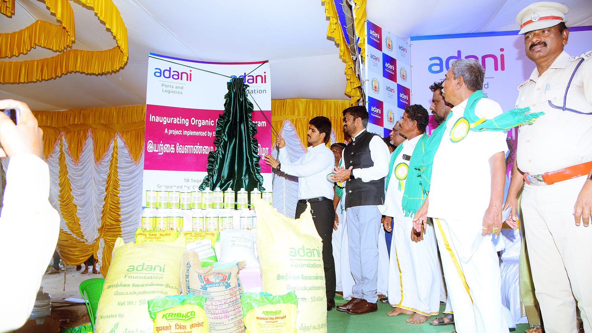 Adani Foundation inaugurates an organic farming program with officials, displaying fertilizers and seeds.