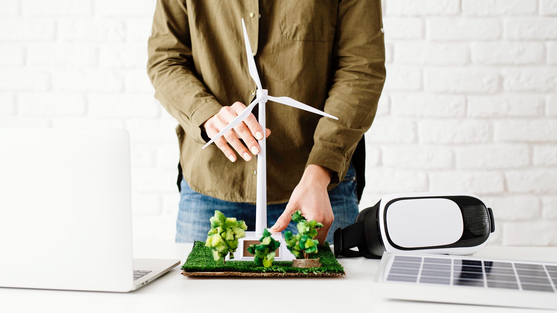 Hands on a wind turbine model with miniature trees, laptop, VR headset, and solar panel.
