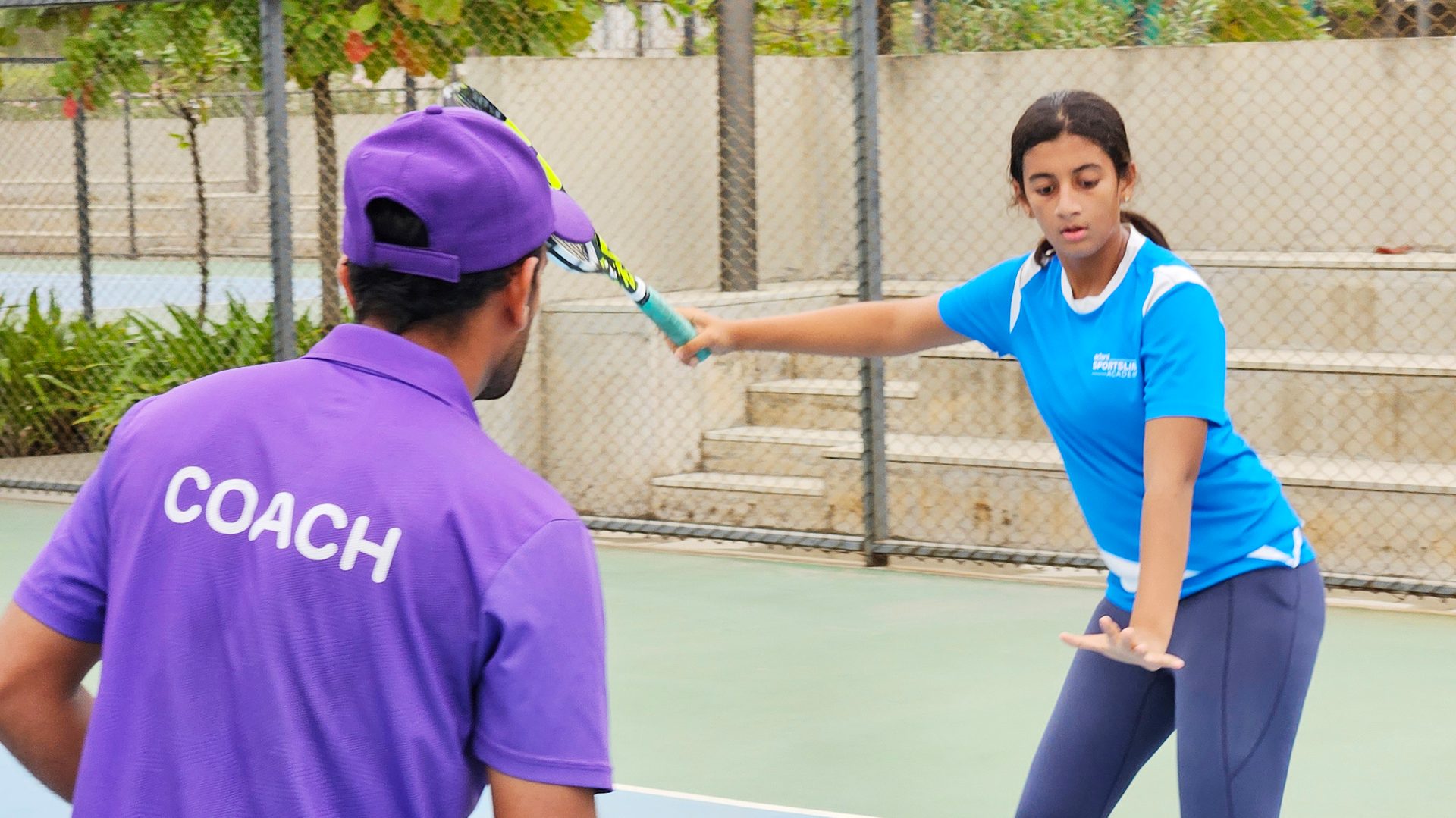 A tennis coach in a purple shirt instructs a girl on a court.