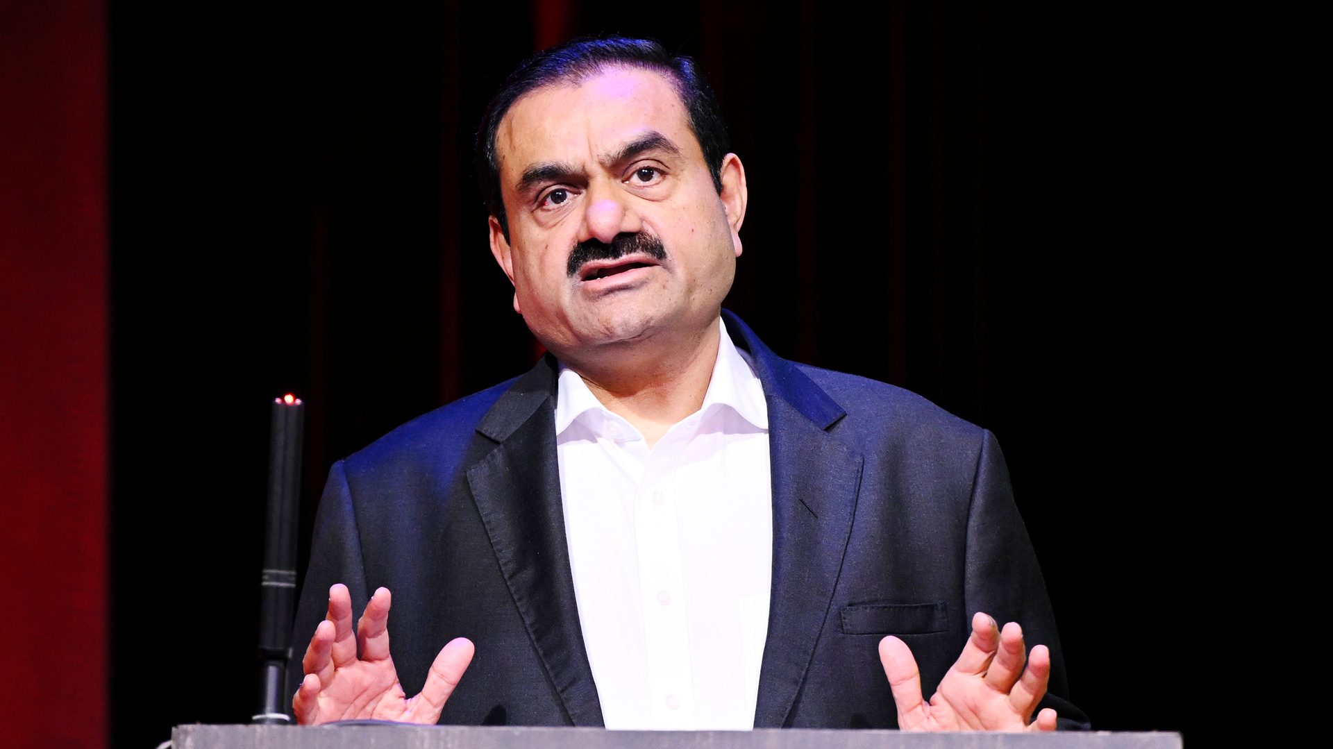 Gautam Adani speaking at a podium, wearing a suit and white shirt, against a dark background.