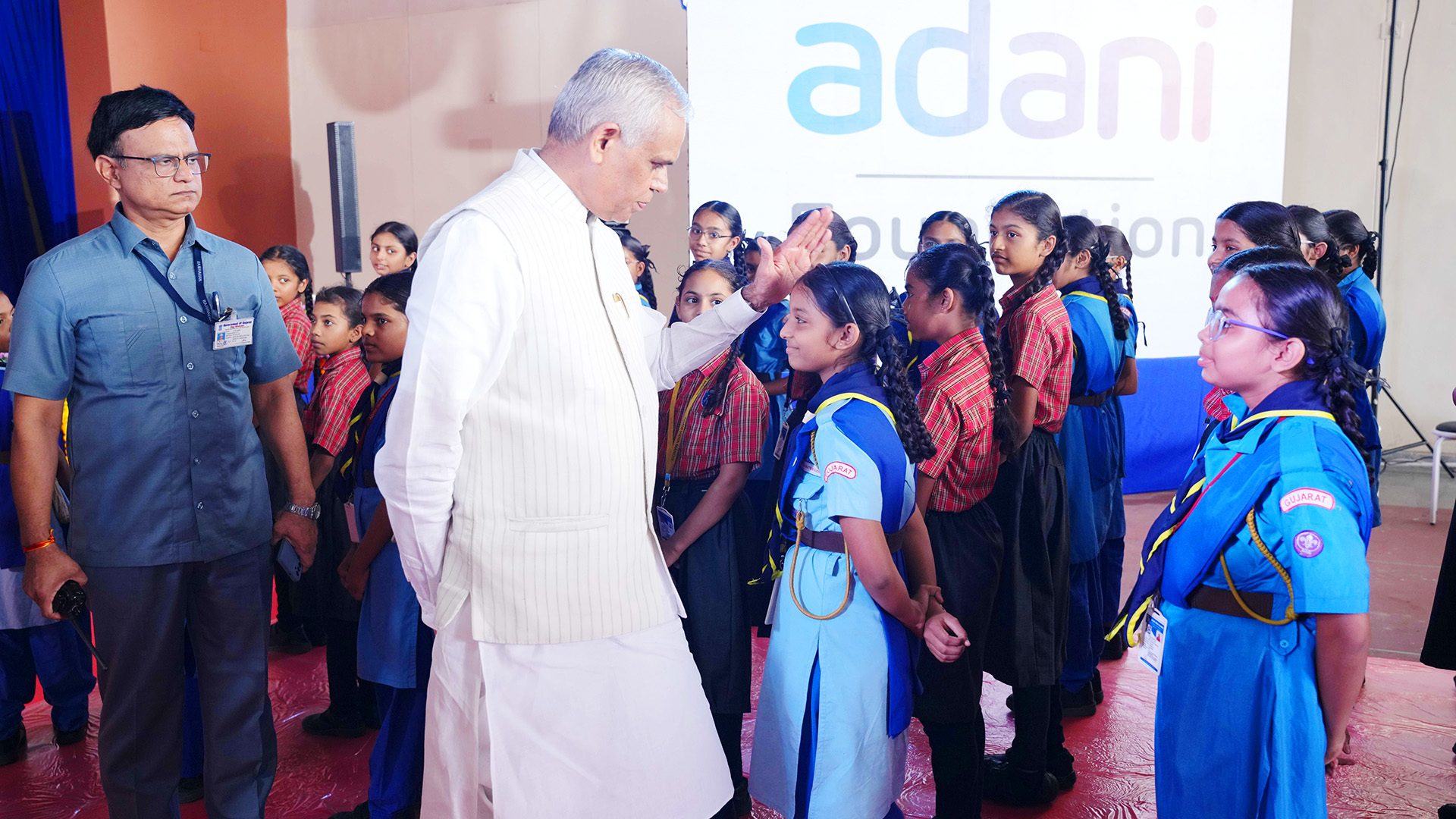 Man in white touches a schoolgirl's head, other girls and 'Adani' logo visible.