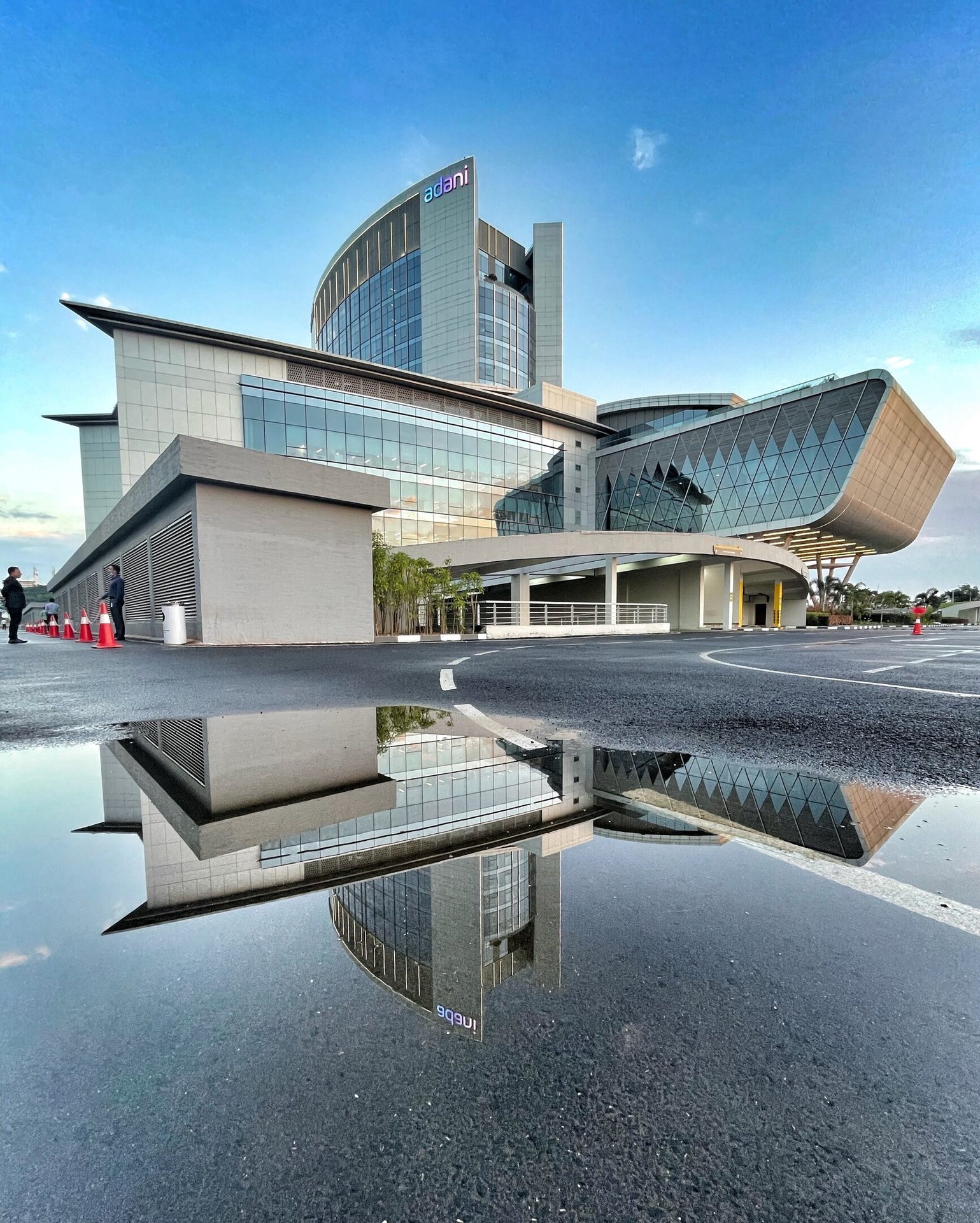 Modern "Adani" building reflected in a large puddle on asphalt under a clear blue sky.