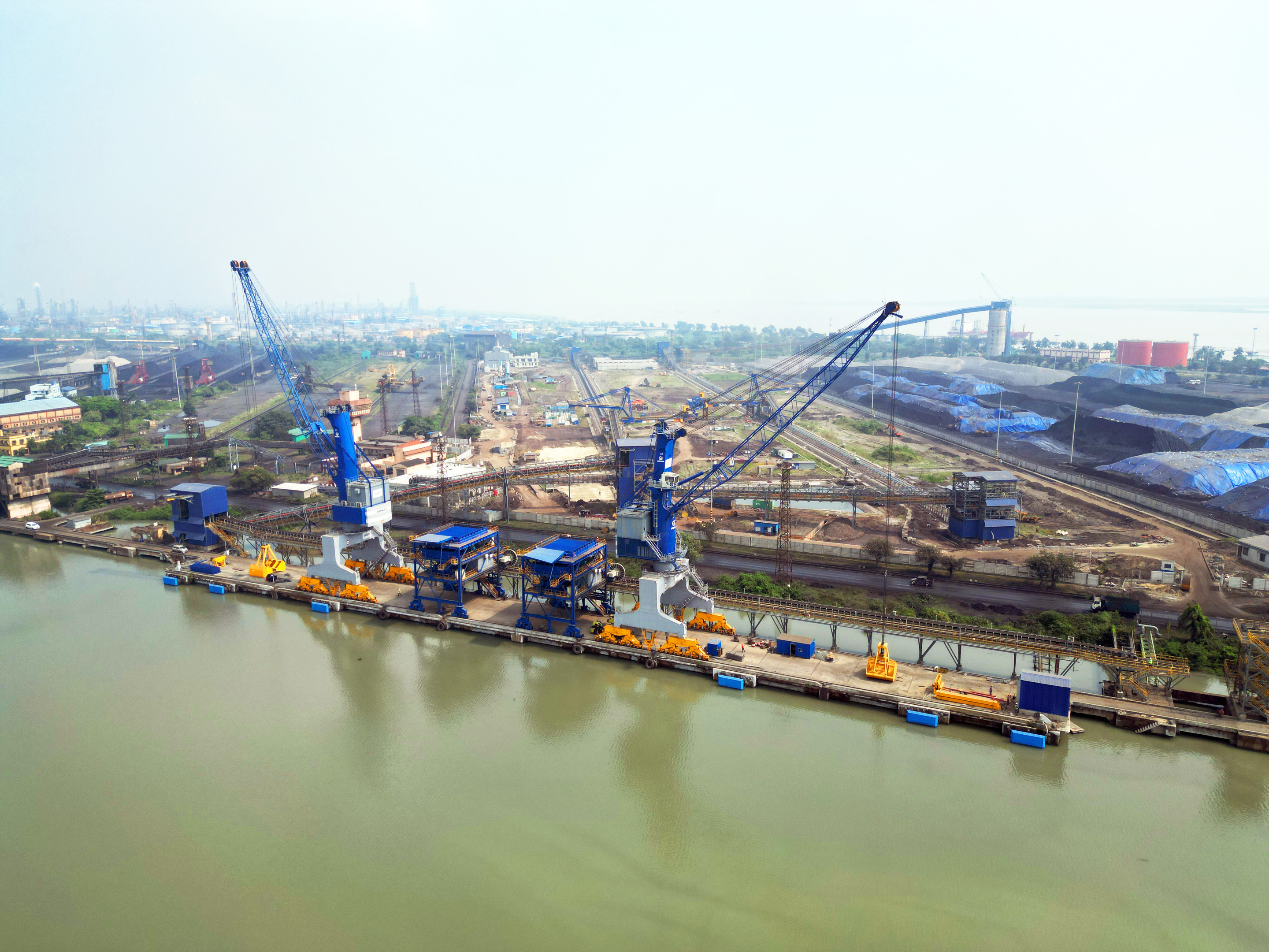 Aerial view of a large industrial port with multiple blue cranes, material piles, and industrial facilities.