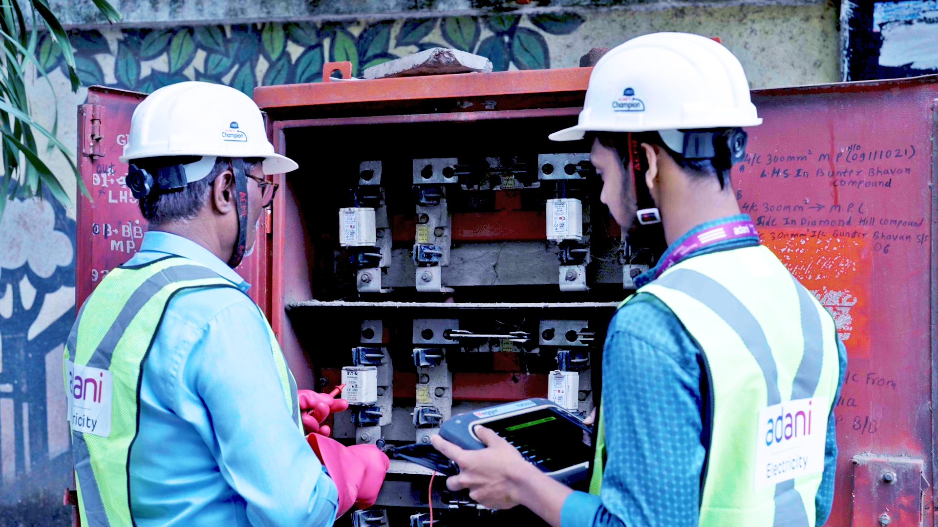 Two electricians in hard hats and safety vests inspect an open electrical panel, one holding a testing device.