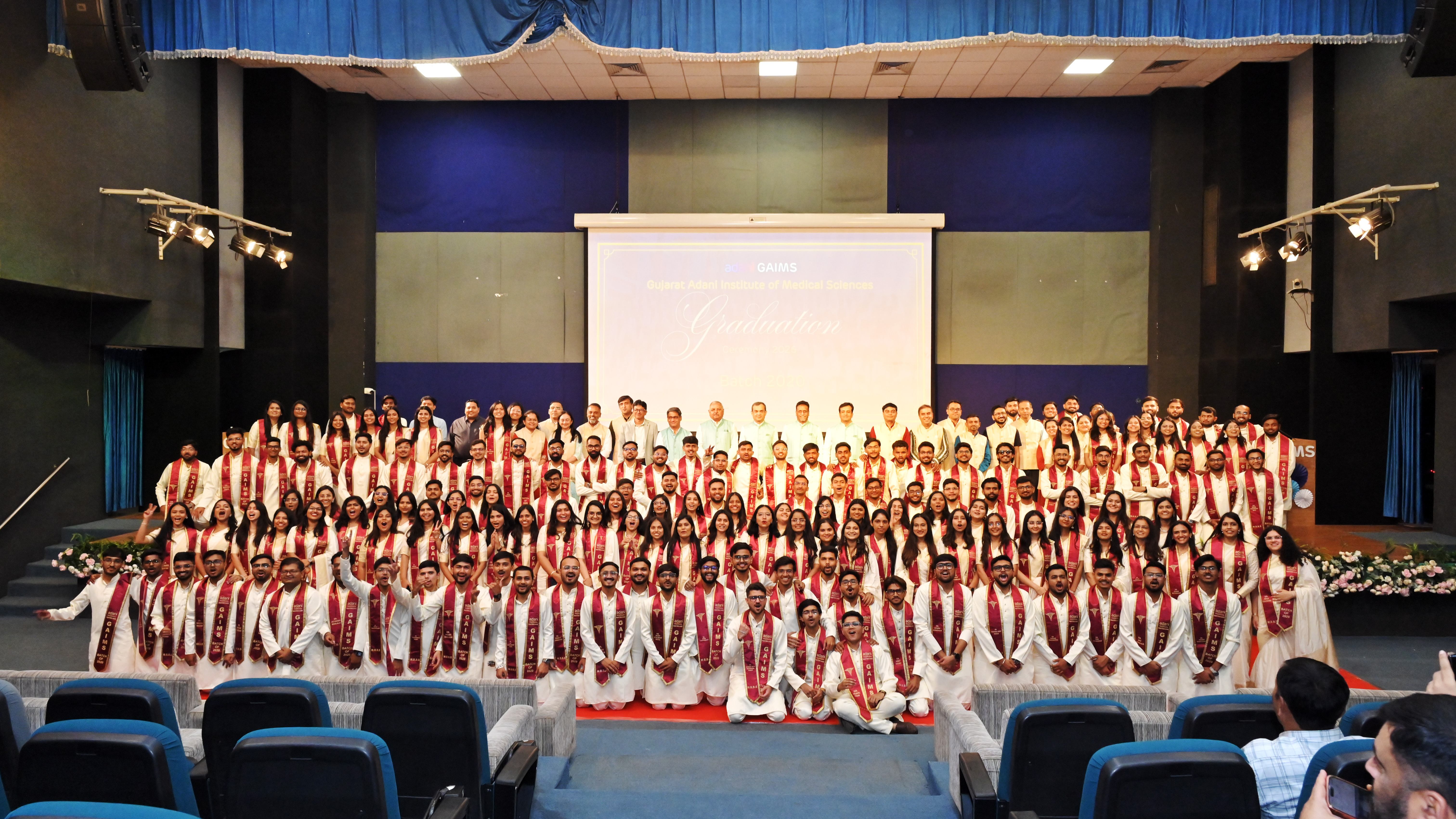 A large group of graduates and faculty in white attire with red stoles posing on stage at a graduation ceremony.
