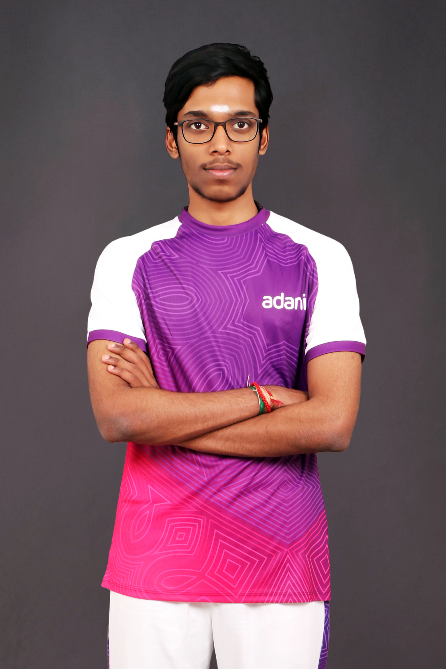 A young man wearing glasses and a purple Adani sports t-shirt with arms crossed, against a dark background.