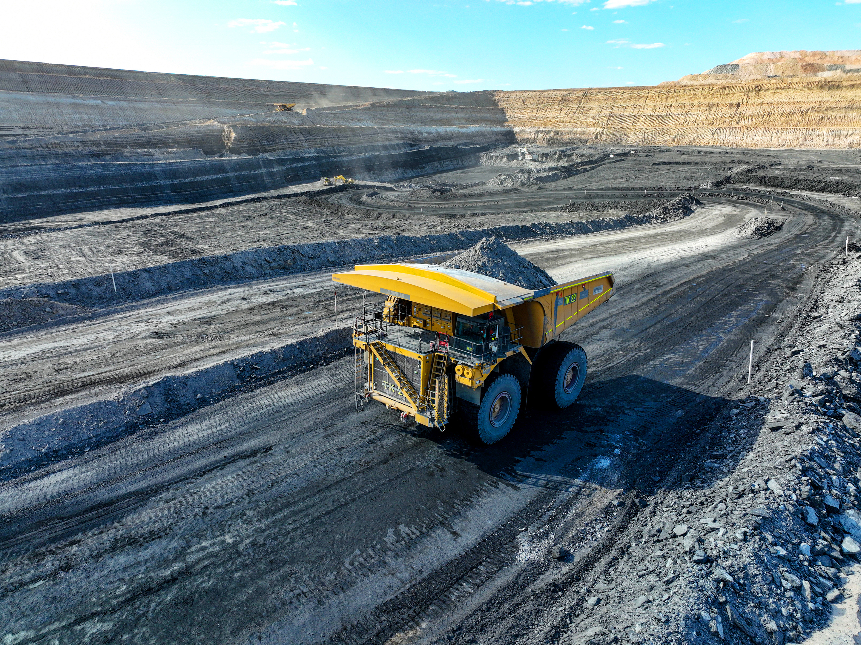 A massive yellow mining dump truck, loaded with material, drives through a large open-pit mine with terraced walls.