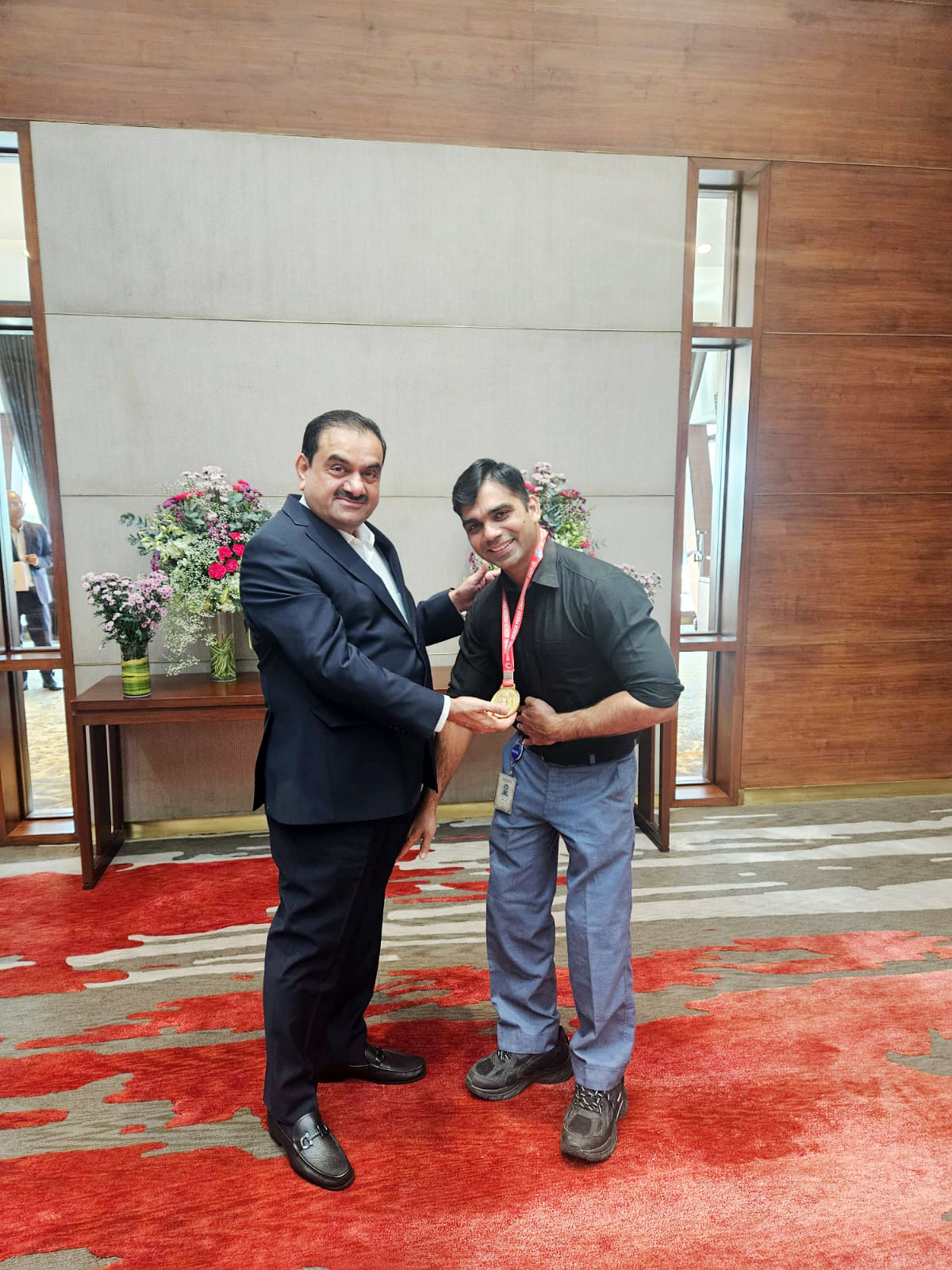 Gautam Adani presenting a gold medal to a man in a black shirt, standing on a red patterned carpet.