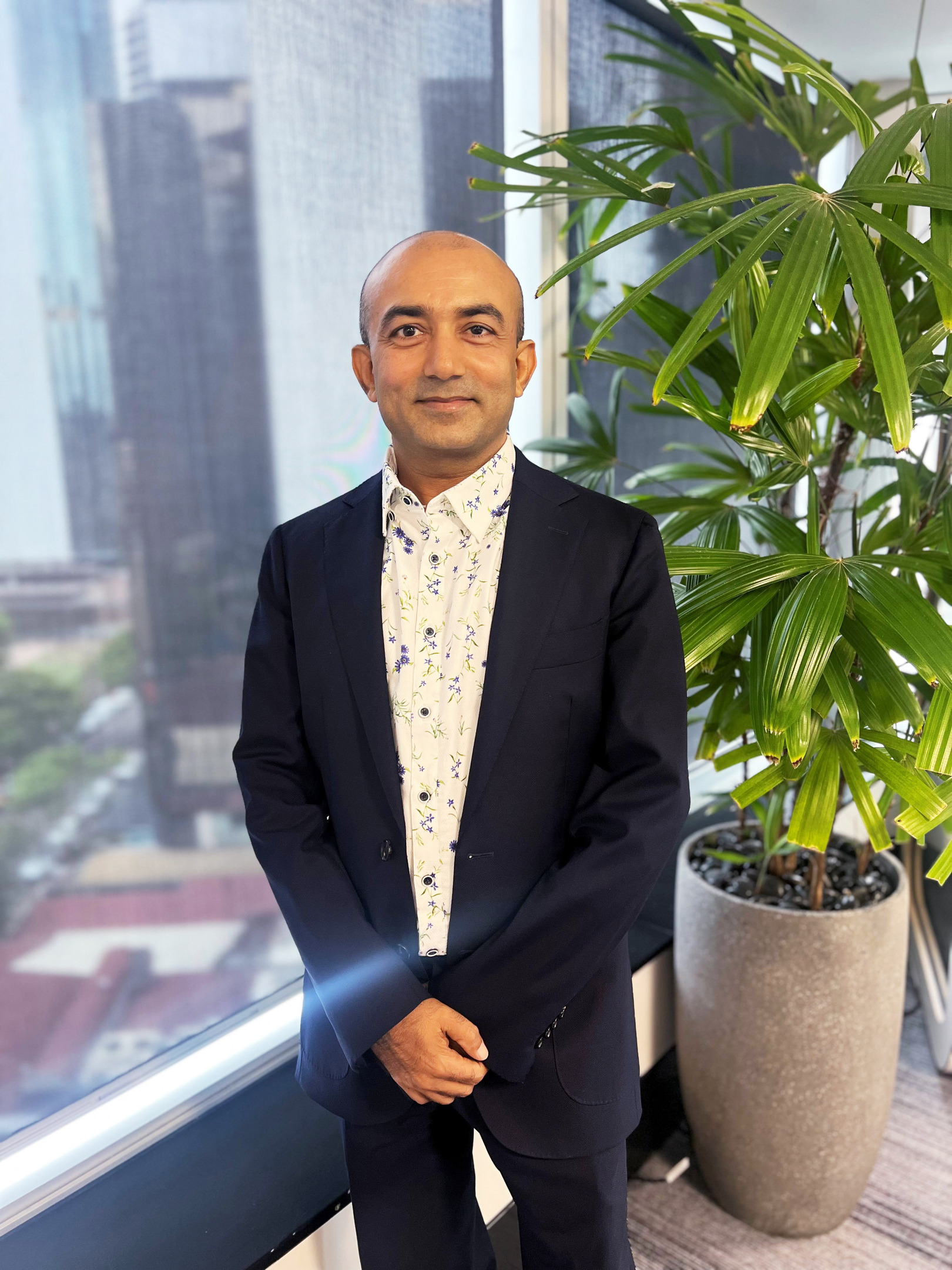 Smiling man in a suit and floral shirt, standing by a window with a city view and a plant.