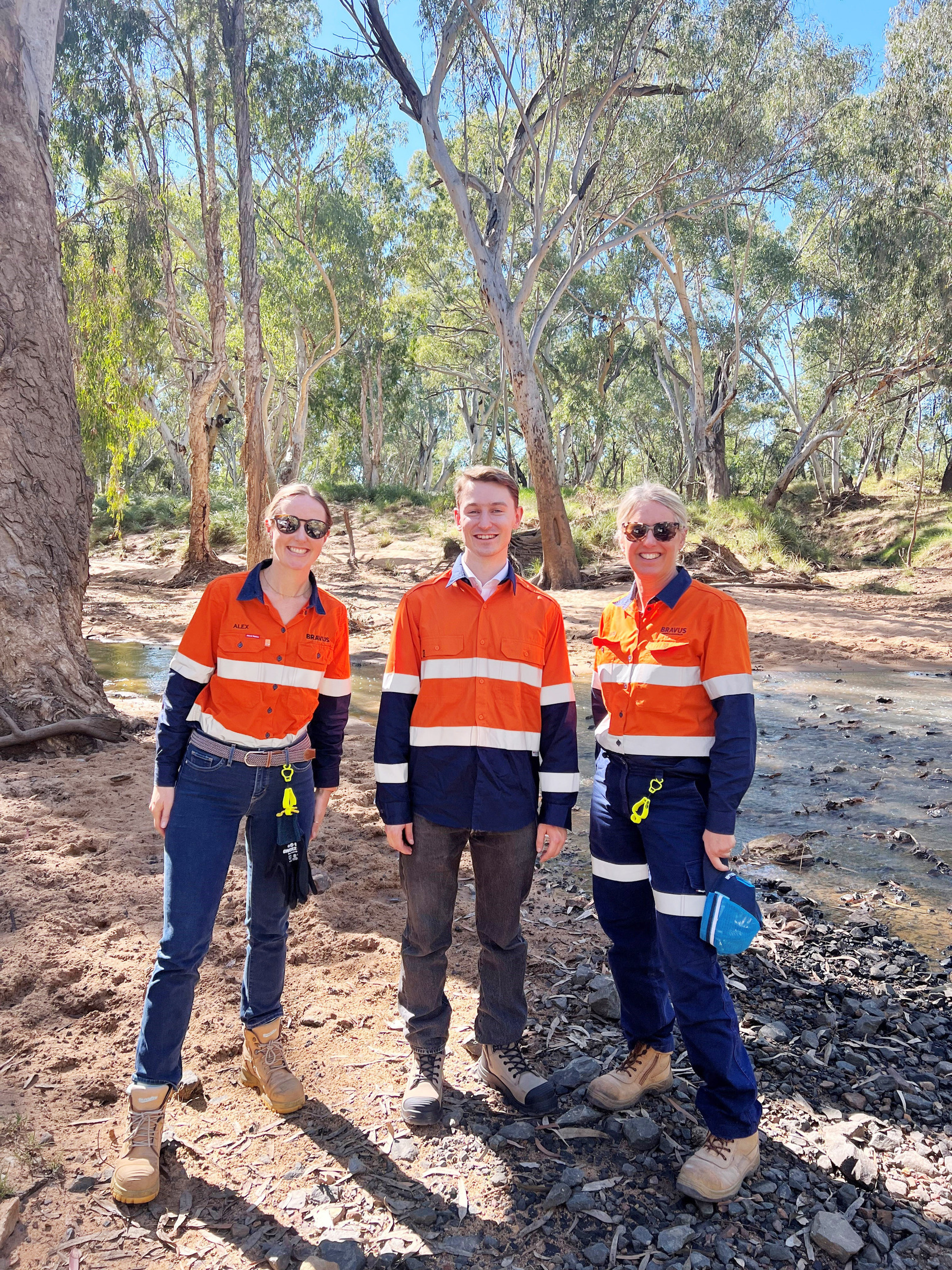 Three individuals in bright orange and navy work attire stand smiling outdoors by a dry creek bed among trees.