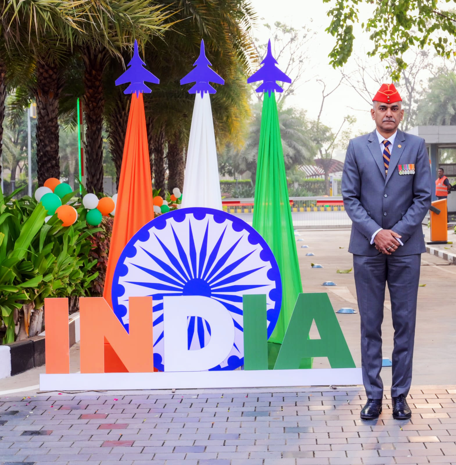 A military officer stands proudly next to a large "INDIA" display with flag colors and fighter jet cutouts.