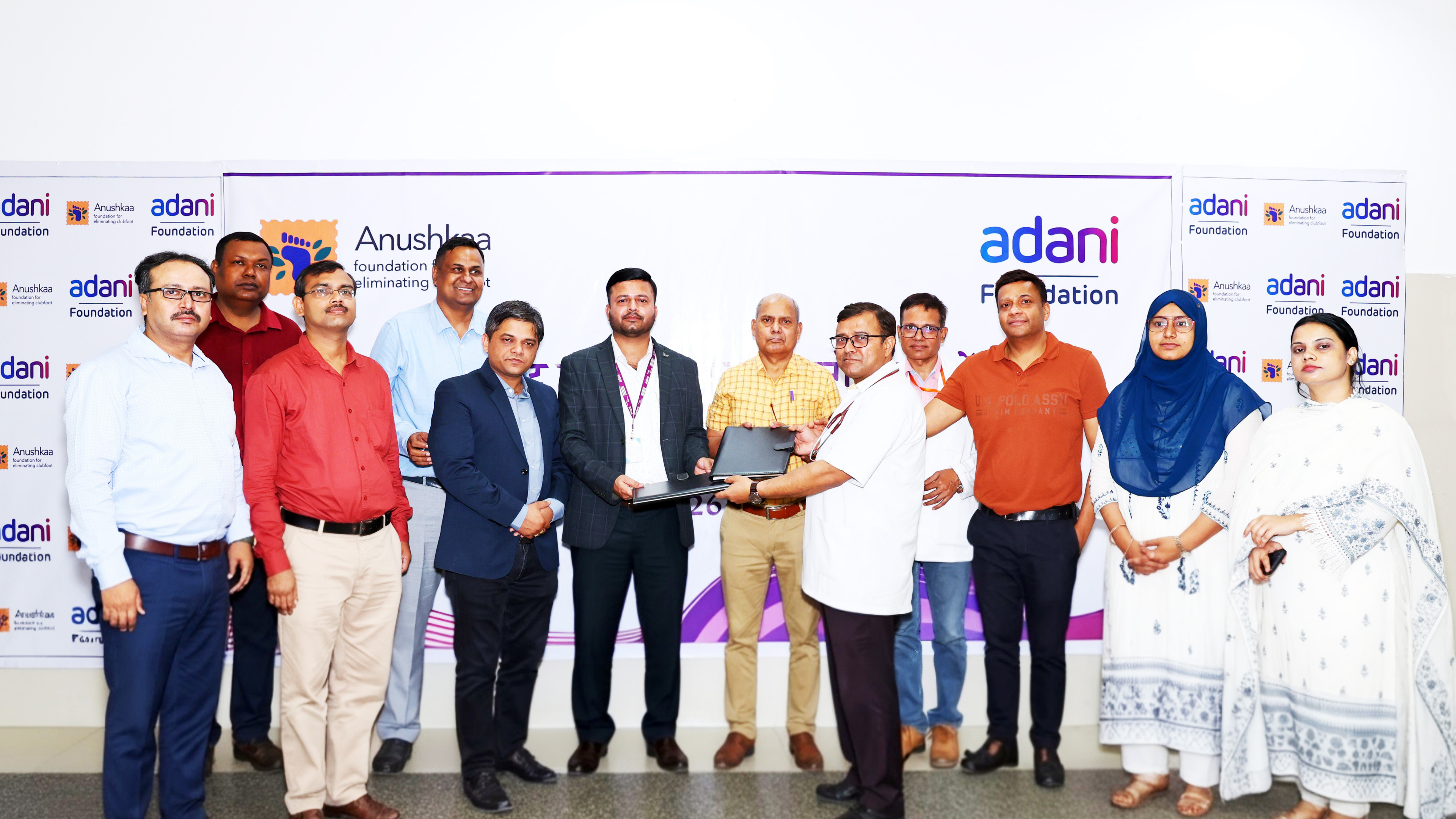 A group of individuals from Adani and Anushkaa foundations gather for an event or signing ceremony, with two men in the center exchanging documents.
